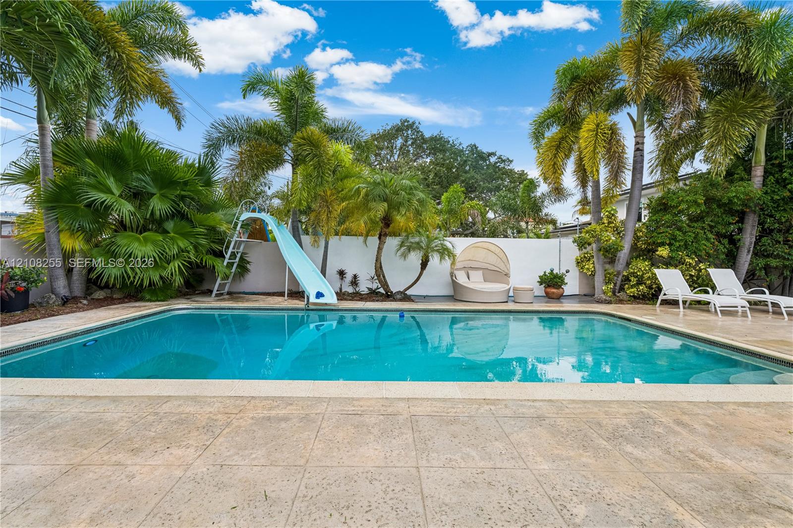 In-ground swimming pool featuring a built-in slide, surrounded by a paved deck and mature palm trees enclosed by a privacy wall