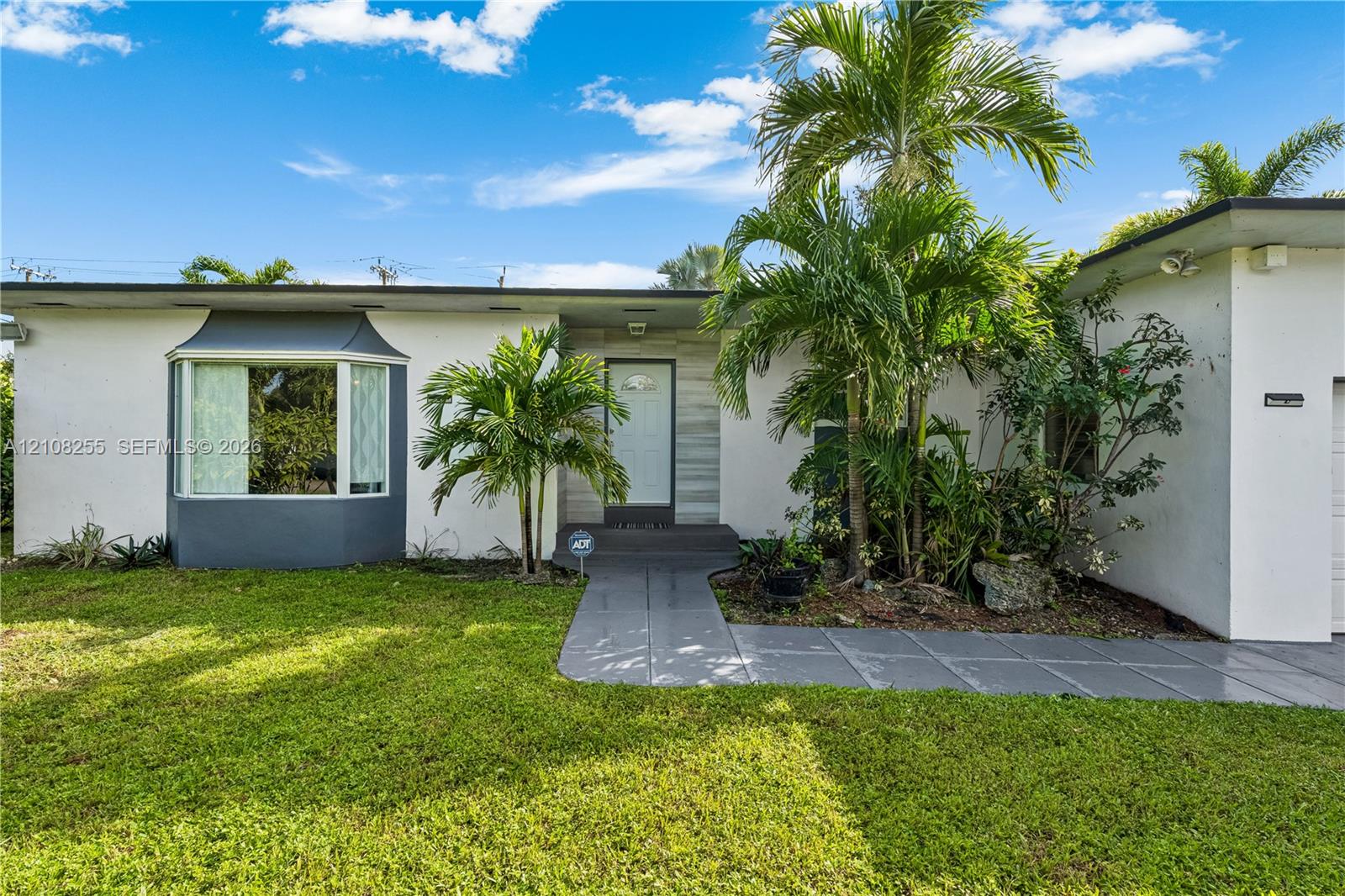 Single-story exterior featuring white stucco finish, front bay window, stone-accented entryway, gray concrete walkway, and established palm landscaping