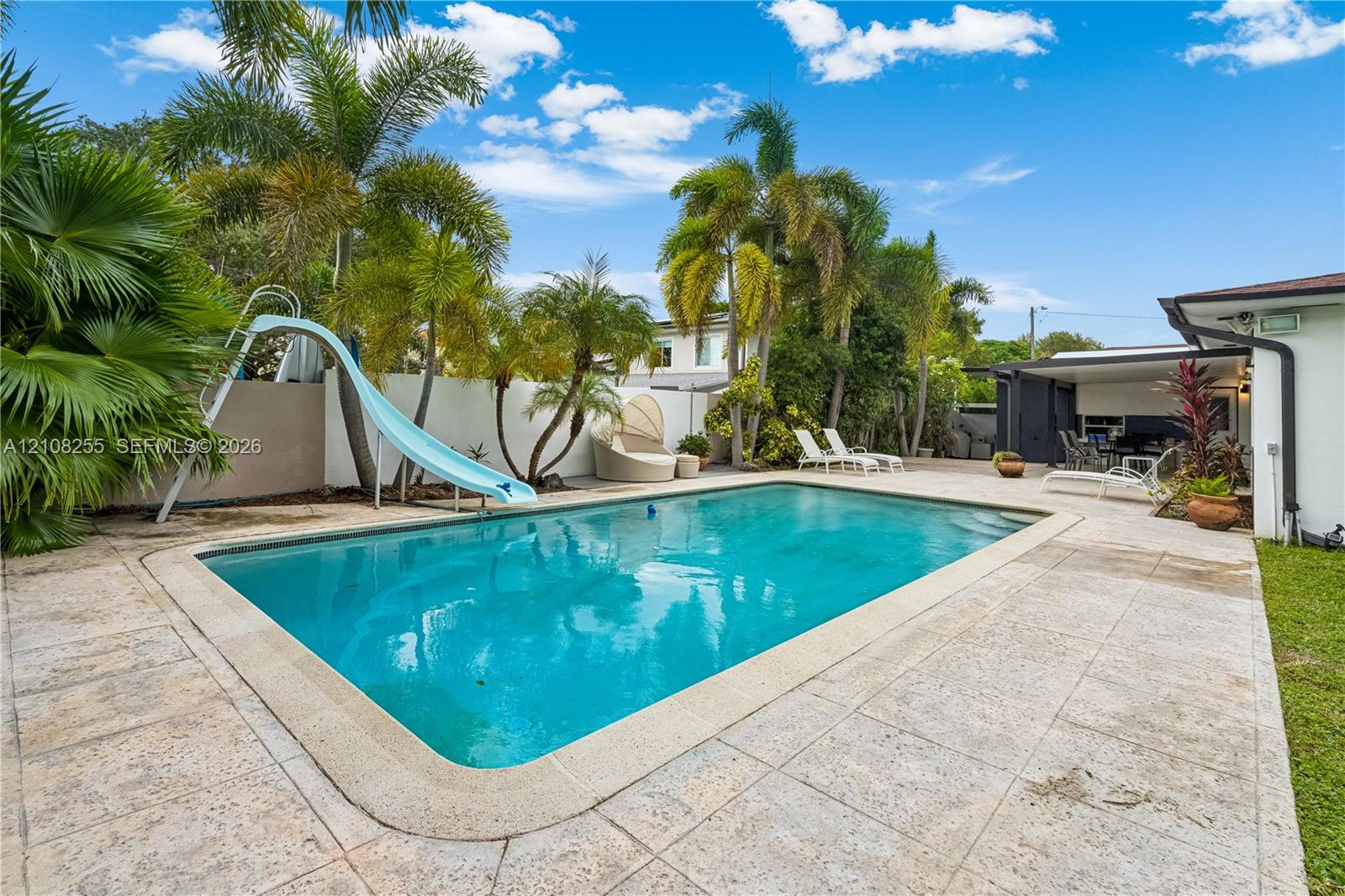 Inground swimming pool with slide, stone-tiled patio, and mature palm trees