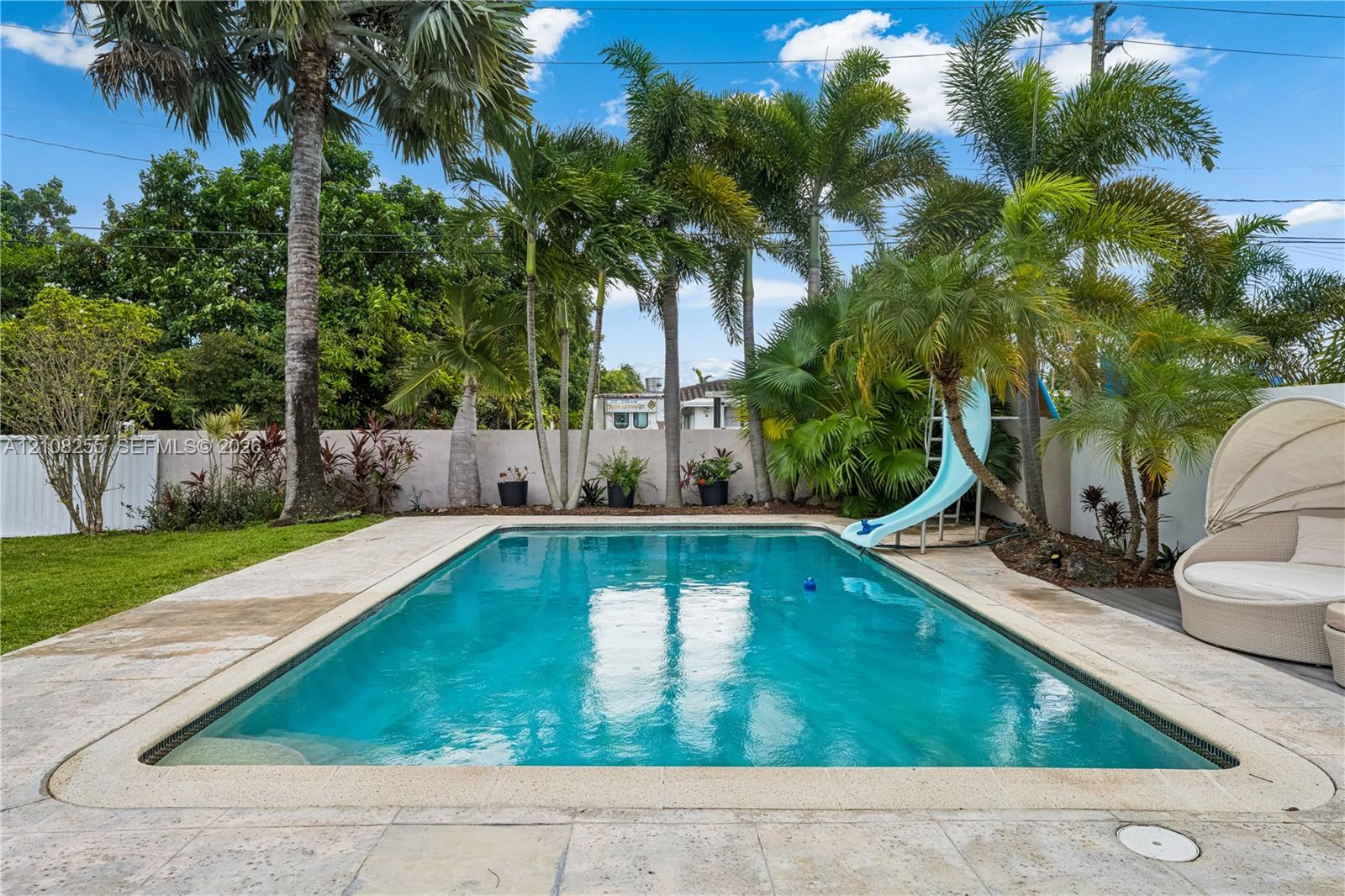 In-ground swimming pool featuring a built-in slide, surrounded by stone paver decking, privacy wall, and mature palm trees