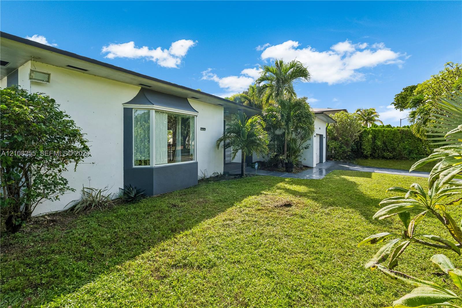 Single-story exterior featuring white stucco siding, a charcoal-trimmed bay window, mature palms, and expansive lawn