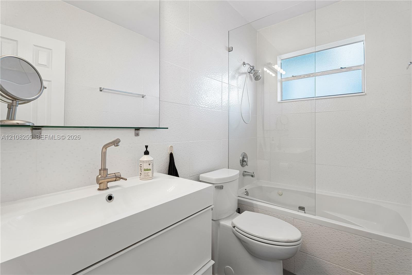 White vanity with an integrated sink, wall-mounted mirror, glass-enclosed shower and tub combination, and brushed-finish metal faucet