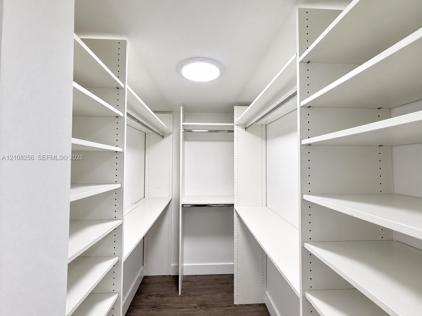 Walk-in storage space featuring floor-to-ceiling white custom shelving, hanging rods, and dark wood-finish flooring