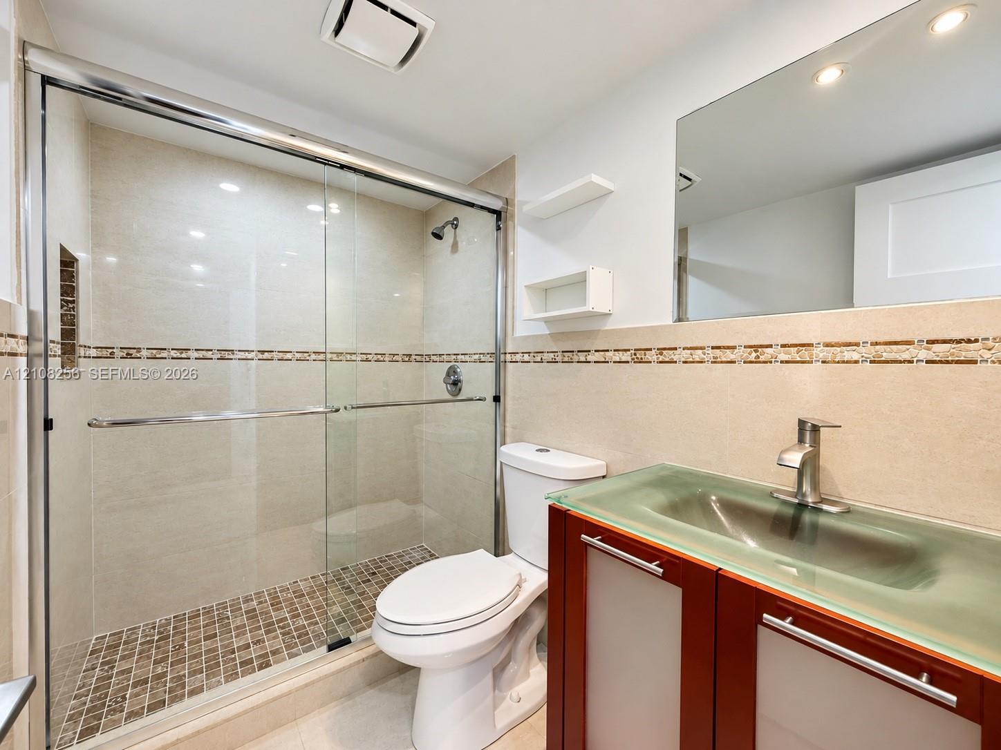 Bathroom featuring a glass-enclosed shower with mosaic tile flooring, a vanity with a frosted glass integrated sink, a large wall-mounted mirror, and decorative tile accents