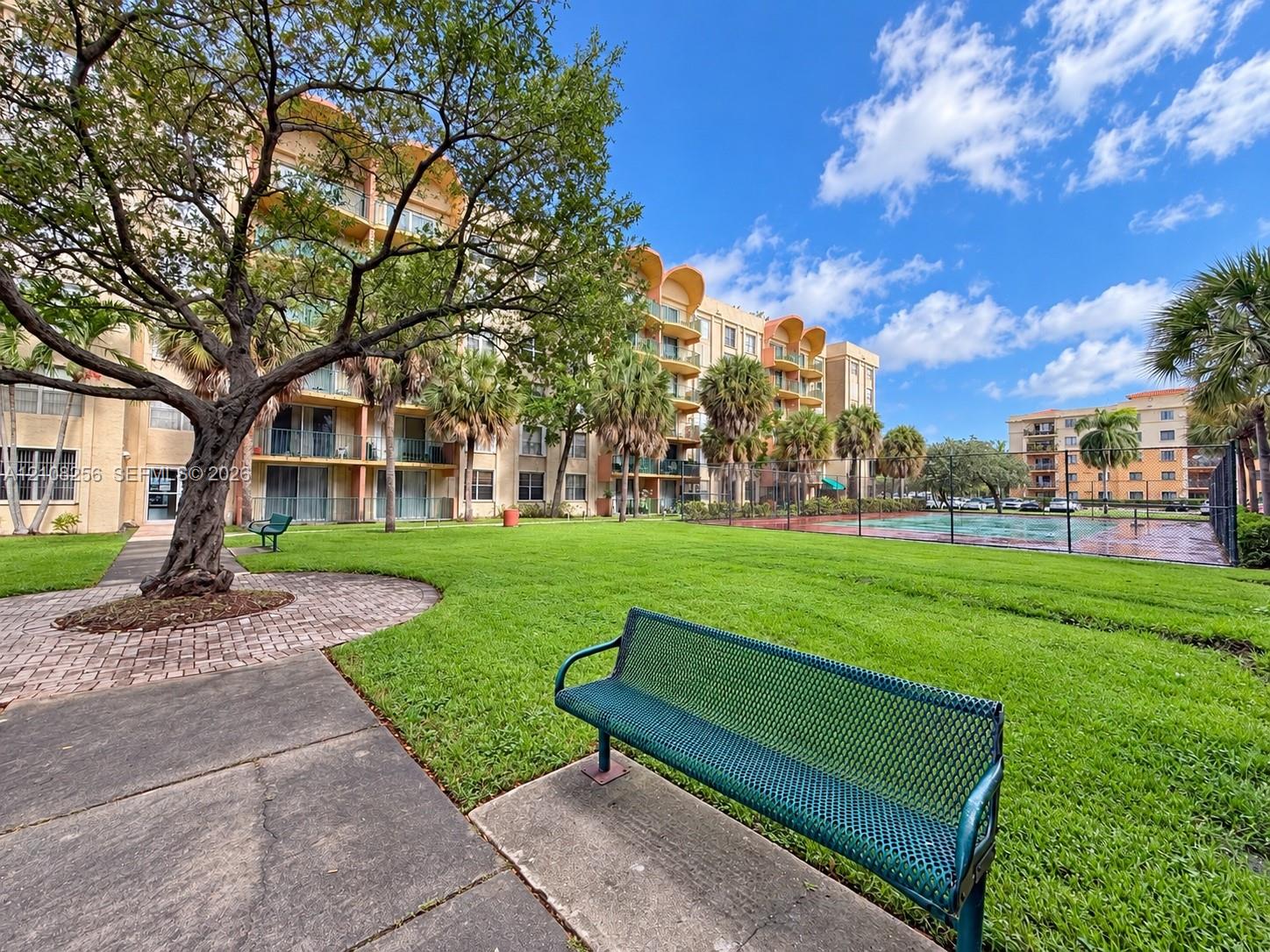 Grounds featuring manicured lawn, mature trees, concrete walking paths, and a fenced recreation court