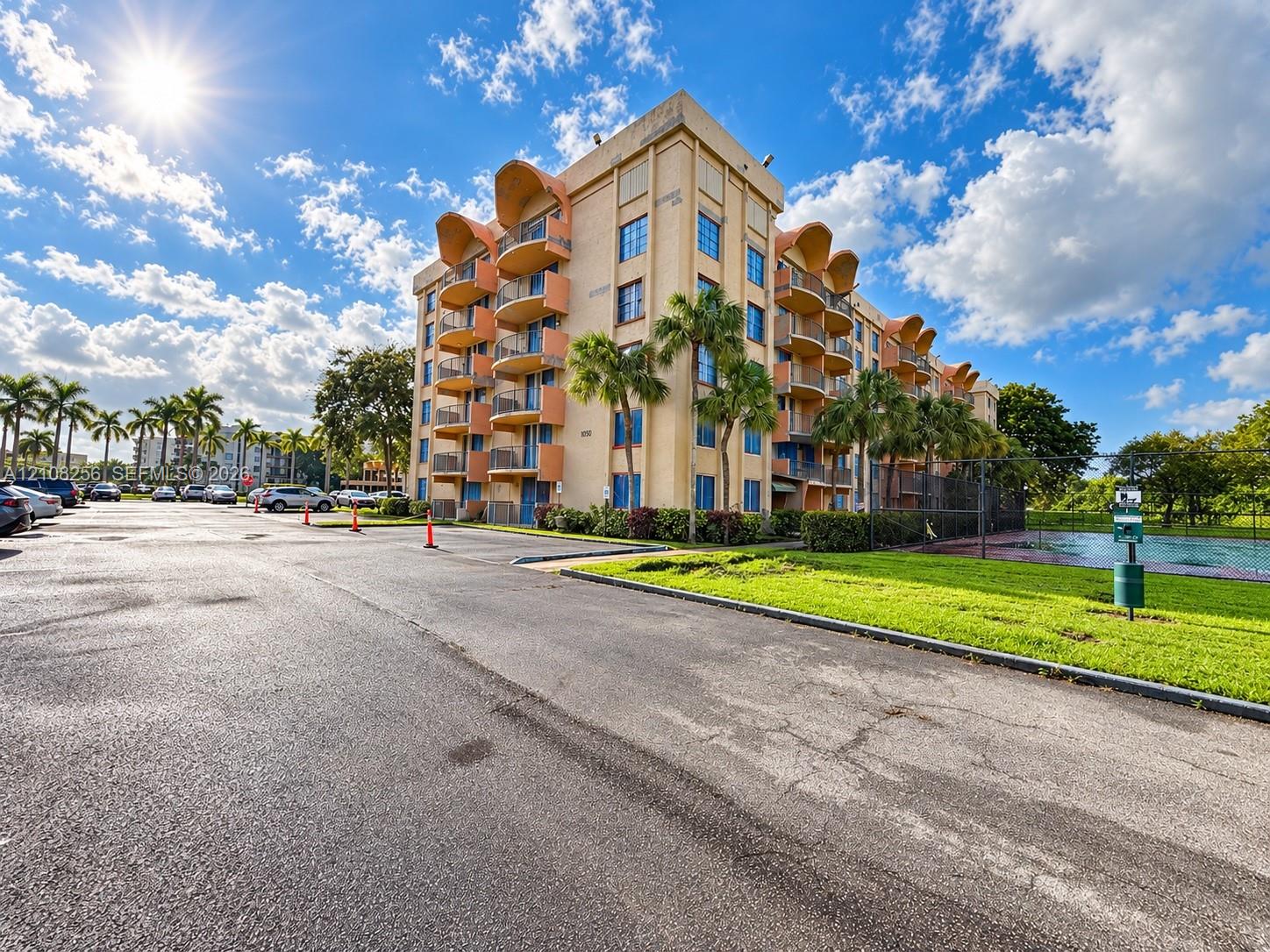 Multi-story stucco exterior featuring arched balconies, metallic railings, paved parking area, and landscaped grounds