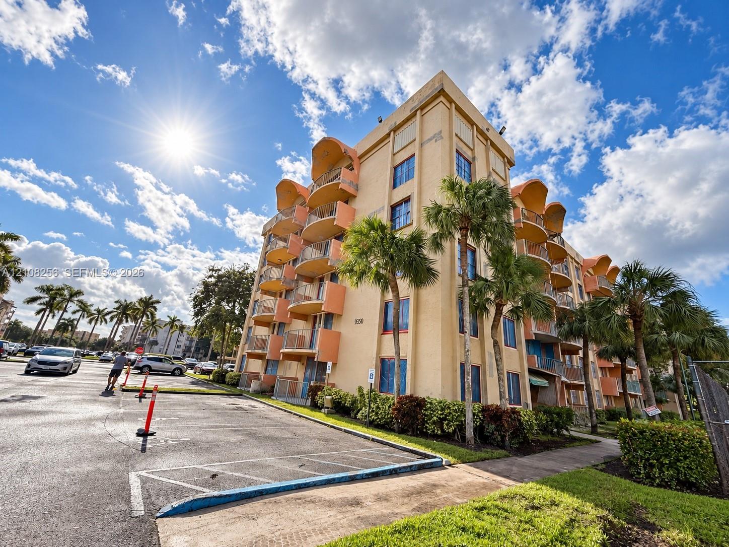 Multi-story building exterior featuring stucco cladding, rounded balconies with railings, and palm tree landscaping