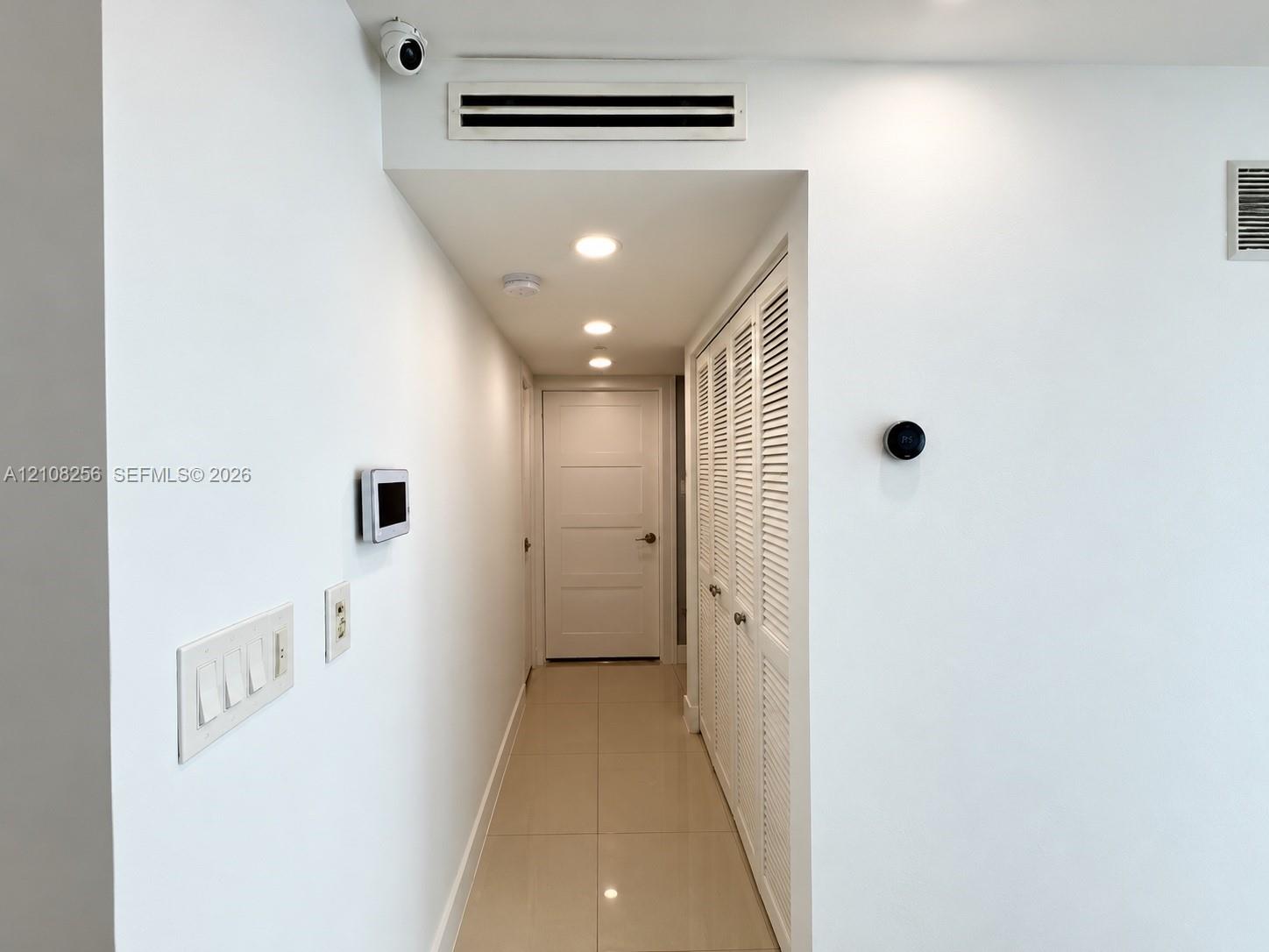Hallway featuring tile flooring, recessed ceiling lighting, louvered closet doors, and integrated smart home wall-mounted controls