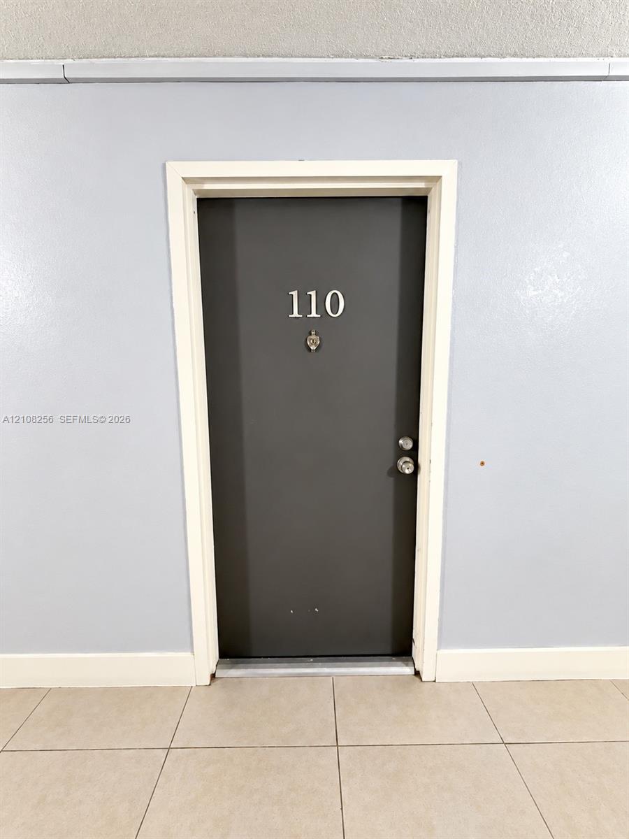 Dark-toned entry door featuring a polished metal door handle, deadbolt lock, and metal kick plate, framed by white trim against light-toned walls and tile flooring