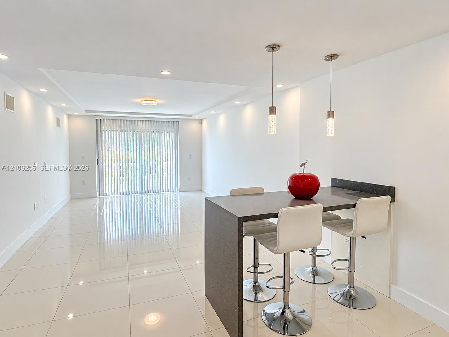 Open-concept interior featuring large-format tile flooring, recessed ceiling lighting, dual cylinder pendant lights, and a waterfall-edge breakfast bar with a solid surface countertop