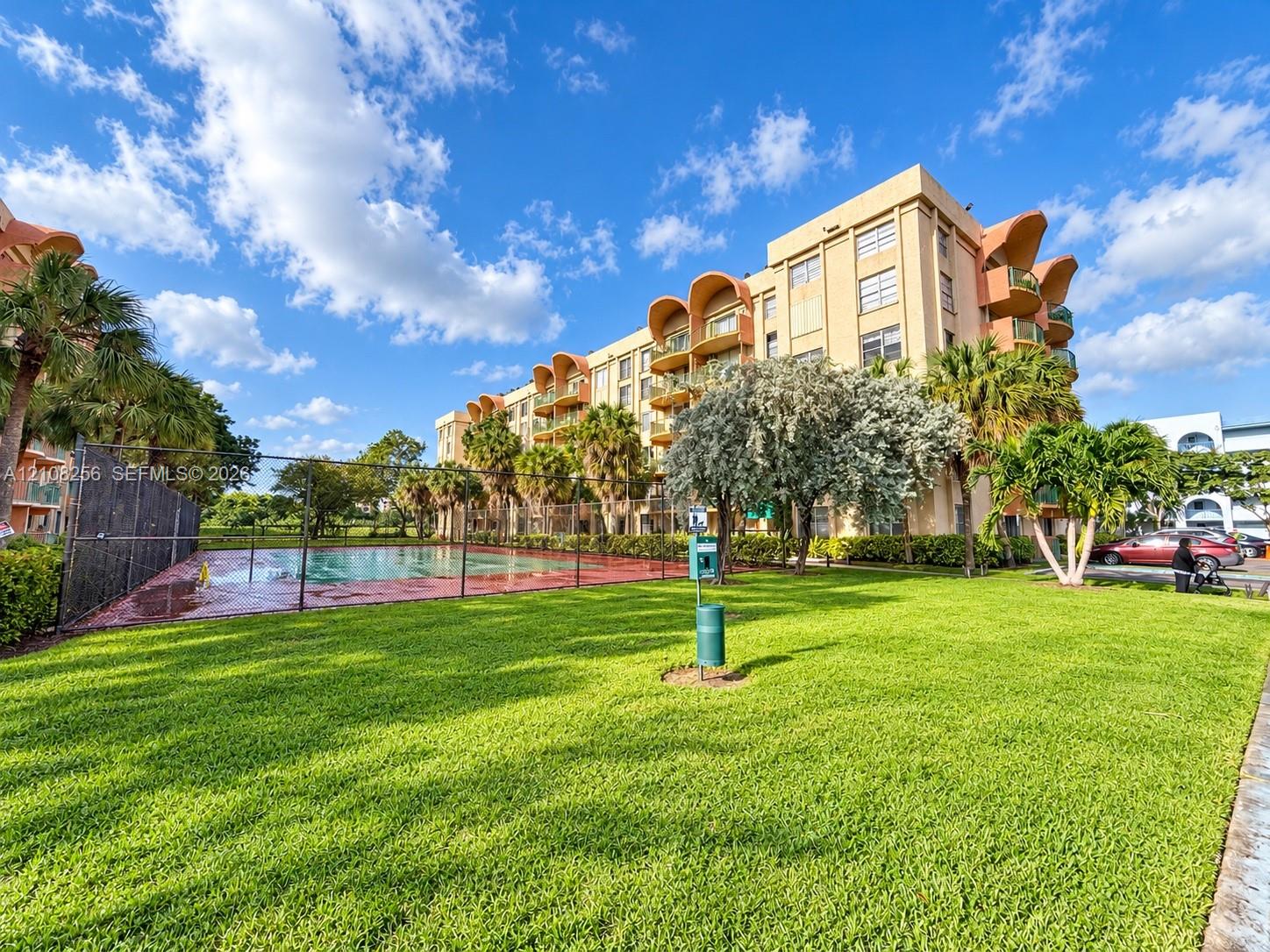 Multi-story building featuring architectural arched balconies, fenced swimming pool, and landscaped green space with mature palm trees