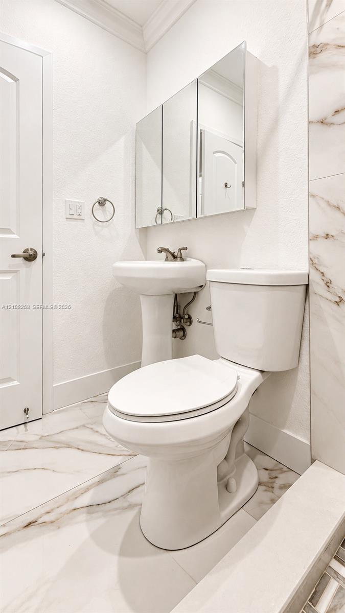 Pedestal sink with metallic faucet, multi-panel mirrored medicine cabinet, elongated toilet, and large-format tile flooring with veined detailing