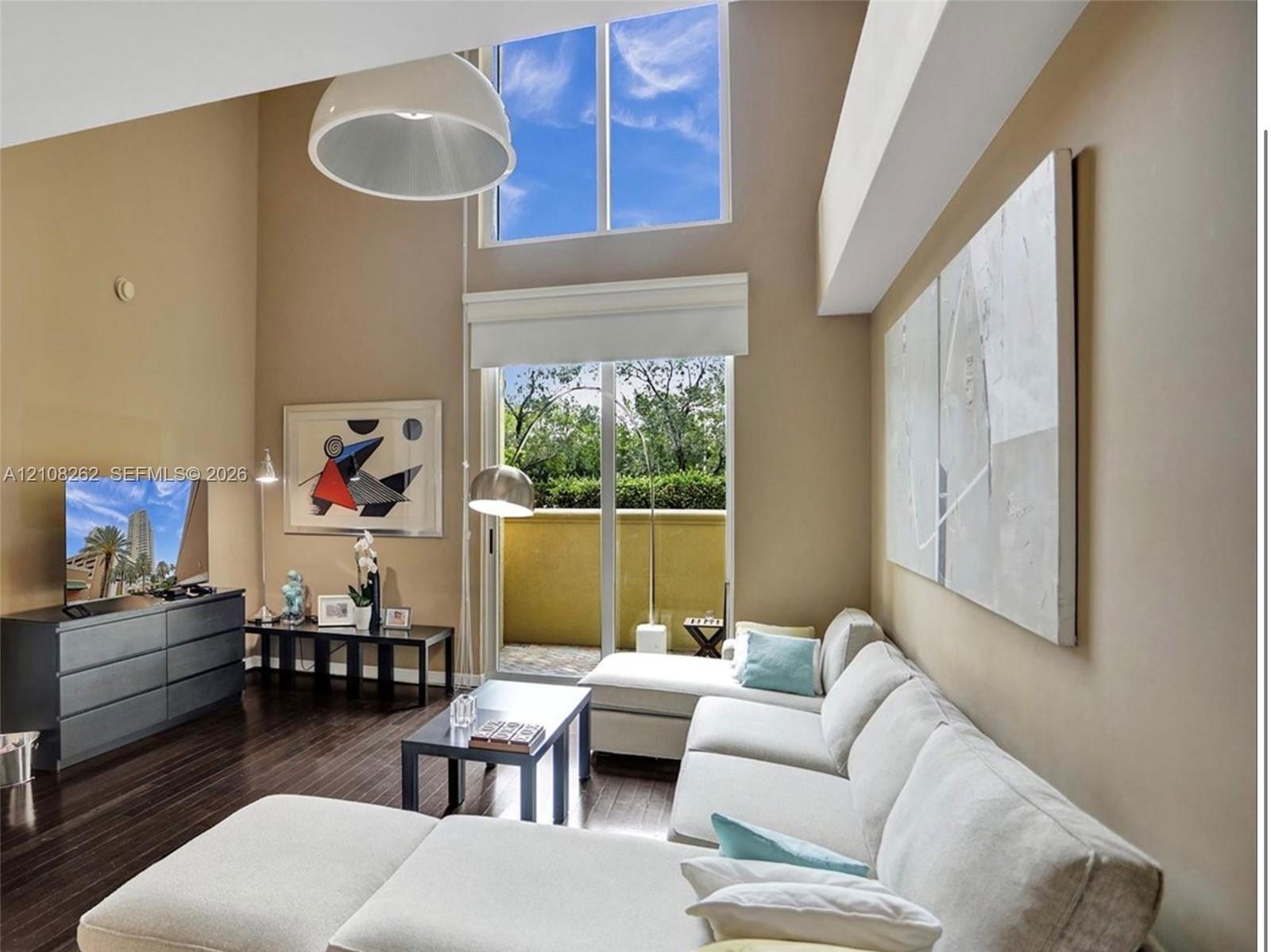 Double-height ceiling, floor-to-ceiling windows, dark wood-finish flooring, and sliding glass doors opening to an exterior patio
