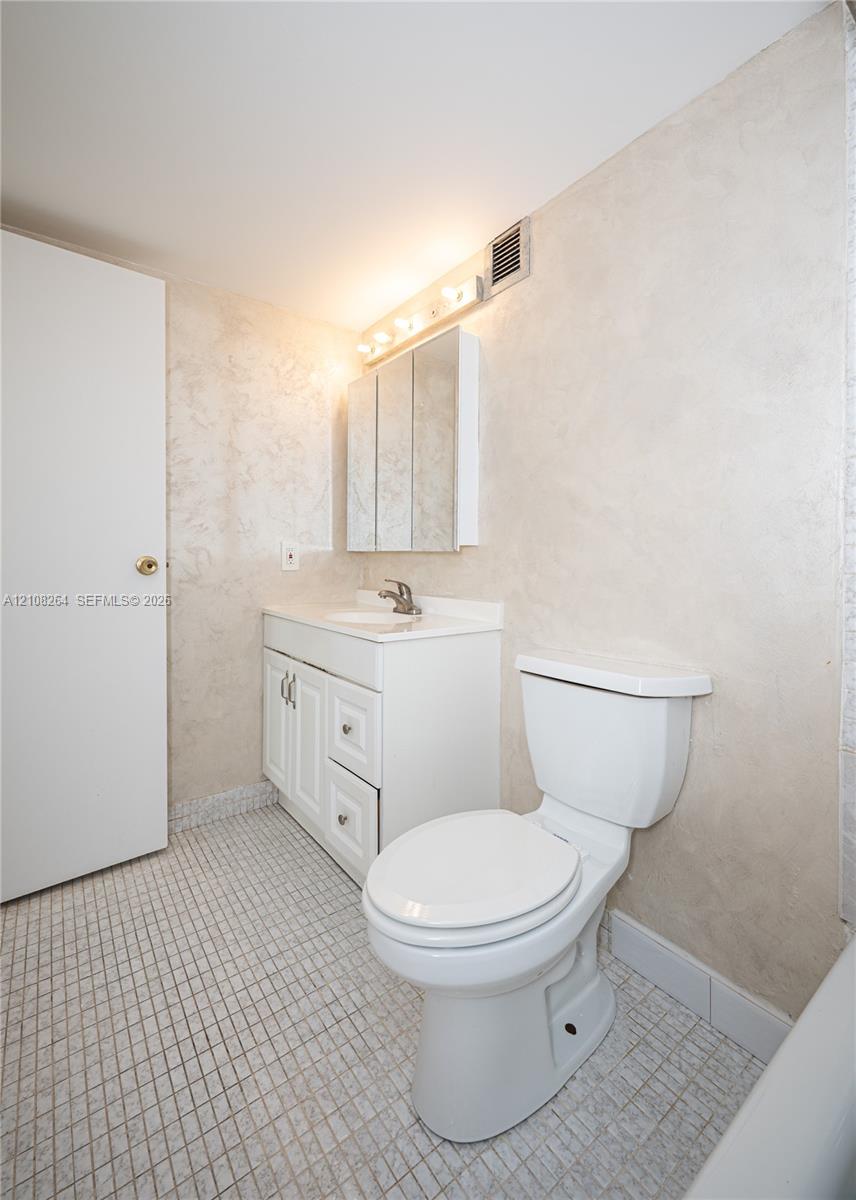 Bathroom featuring white vanity cabinetry with integrated countertop, tri-fold medicine cabinet, wall-mounted light fixture, and ceramic tile flooring