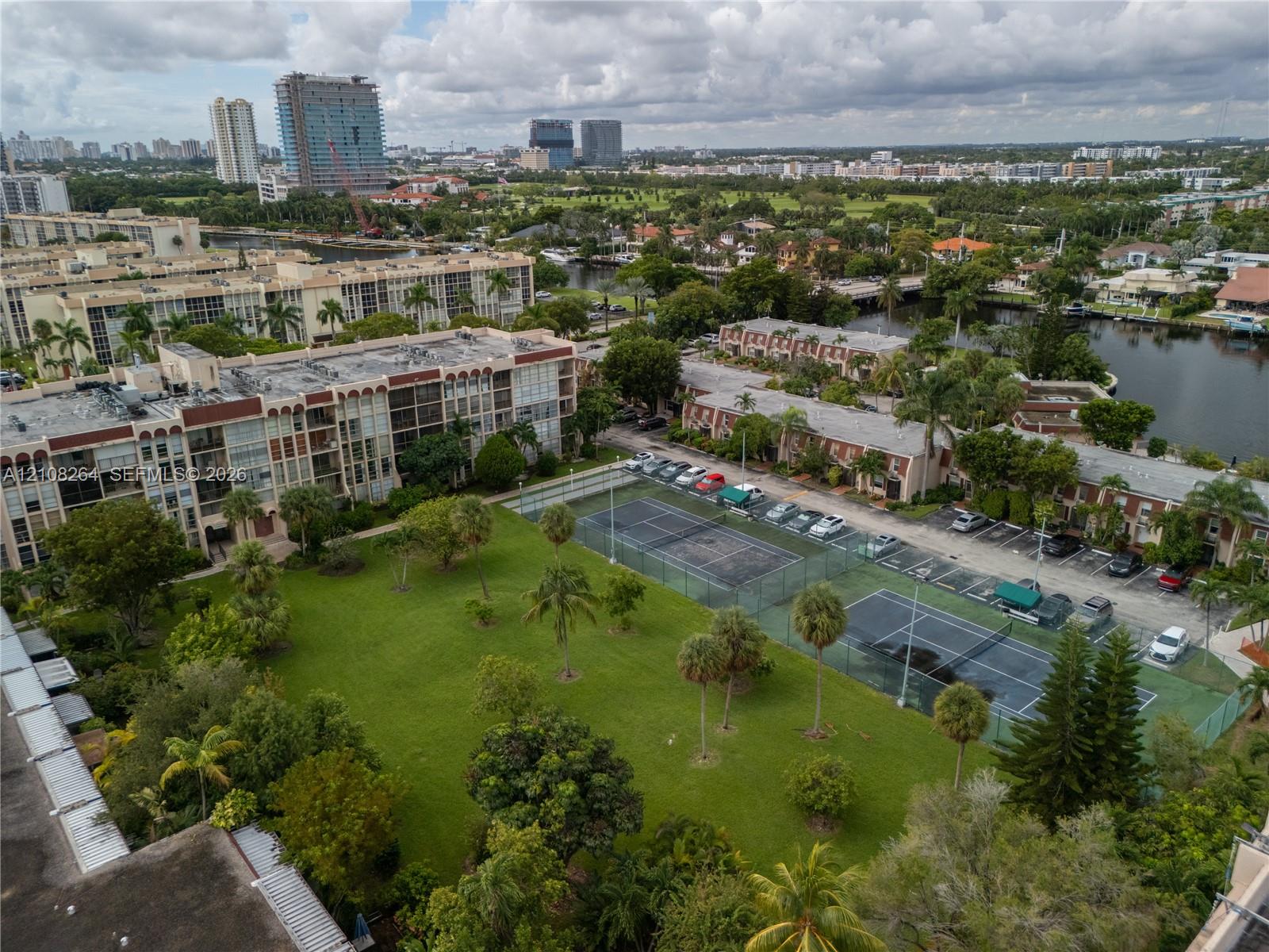 Aerial perspective of community grounds featuring two tennis courts, open green space, mature palm trees, and canal frontage
