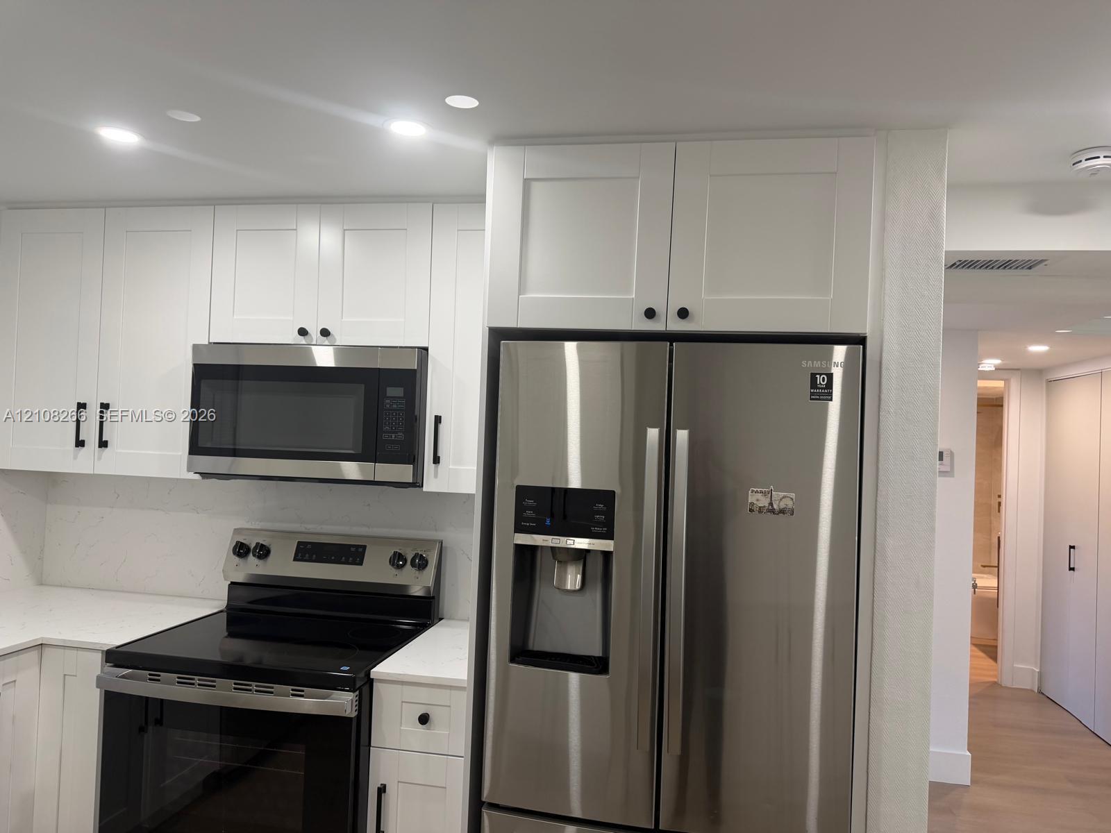 White shaker-style cabinetry with matte black hardware, stone-finish countertops, stainless steel side-by-side refrigerator, built-in stainless steel microwave, and recessed lighting
