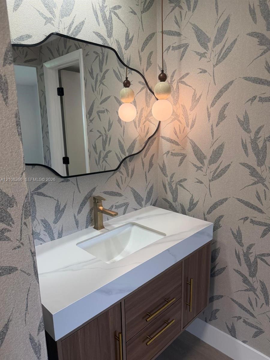 Floating vanity with wood-finish cabinetry, white countertop, integrated basin, brushed gold-tone hardware, and sculptural pendant lighting set against patterned wall covering with a black-framed accent mirror