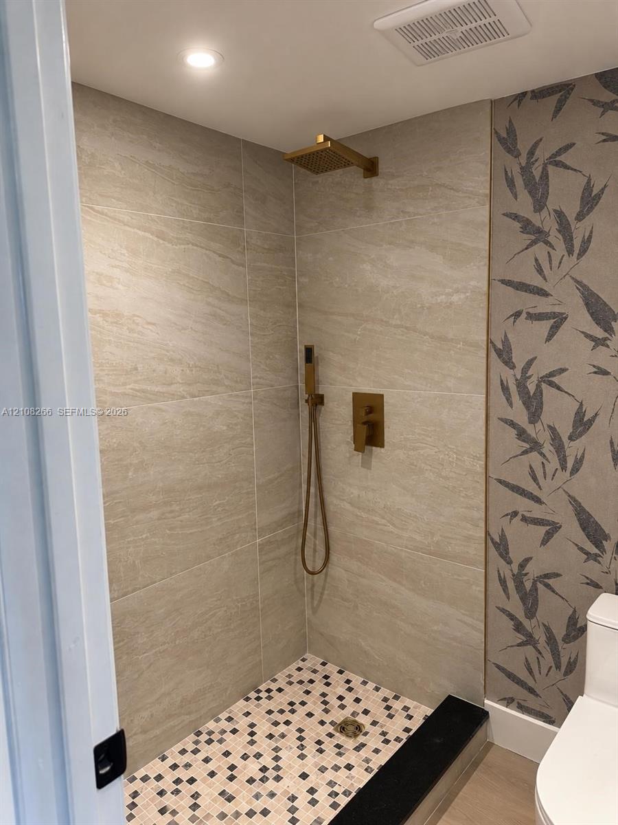 Walk-in shower featuring large-format stone-finish tile surround, mosaic floor tiling, gold-tone rainfall showerhead, and matching hand-held sprayer