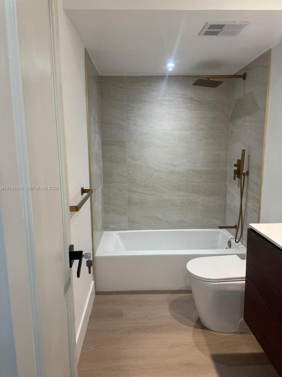 Bathroom featuring a soaking tub, floor-to-ceiling tile surround, metallic finish shower fixtures with handheld sprayer, wood-finish vanity, and wood-finish flooring