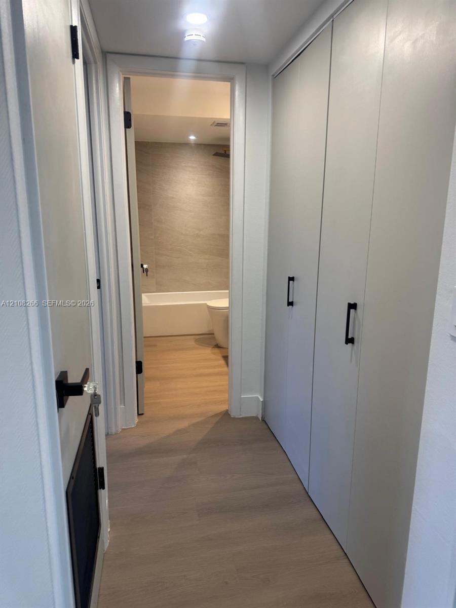 Hallway featuring floor-to-ceiling built-in cabinetry with matte black hardware, wood-finish flooring, recessed lighting, and access to a bathroom with a tiled wall surround and tub