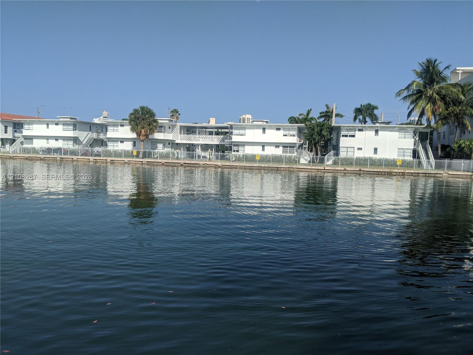 Multi-story exterior featuring white stucco facade, exterior staircases, metal fencing, and established palm trees situated along the water's edge
