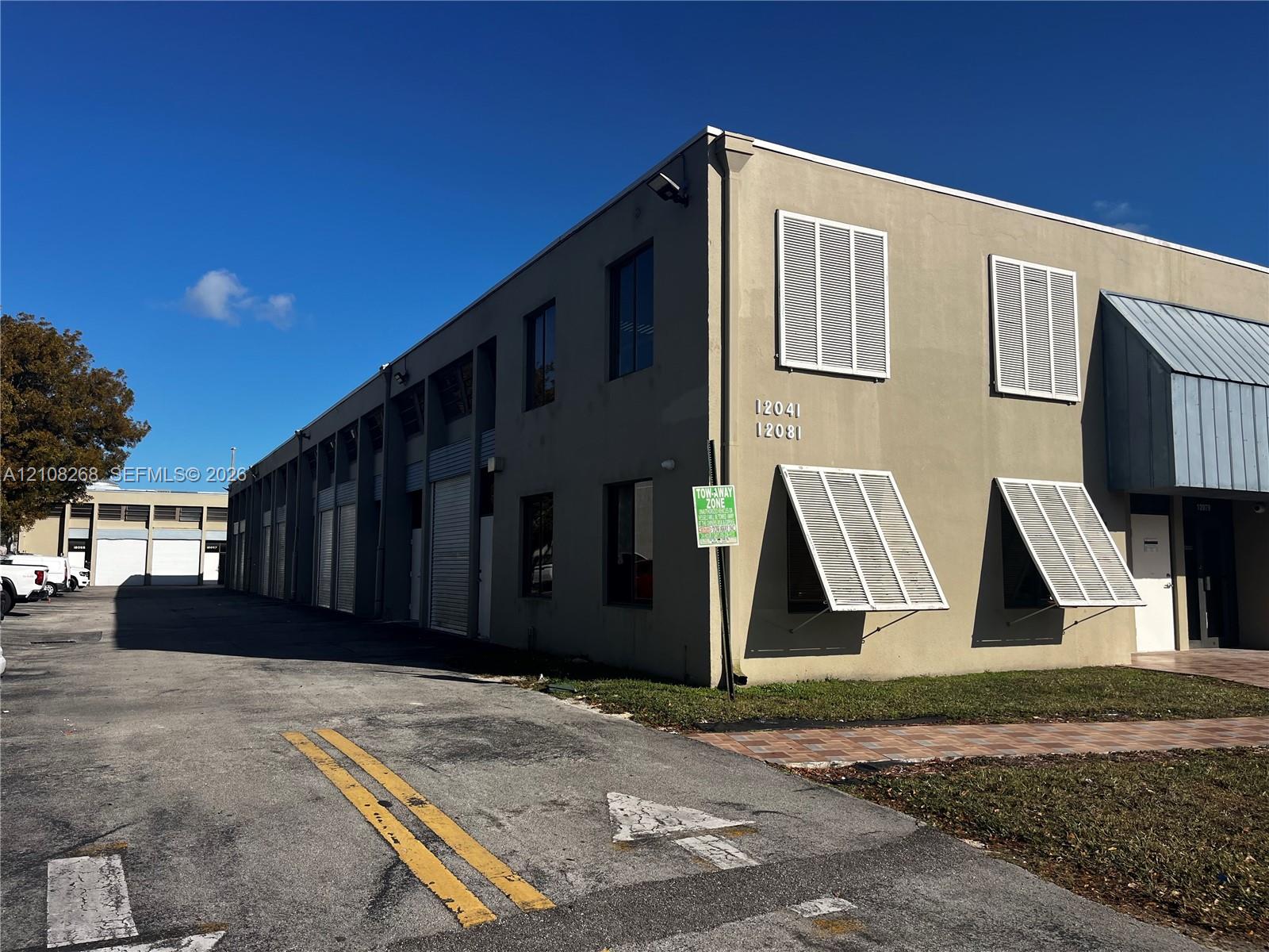Multi-unit industrial building exterior featuring a concrete facade, metal roll-up doors, exterior shutter accents, and an expansive asphalt parking area