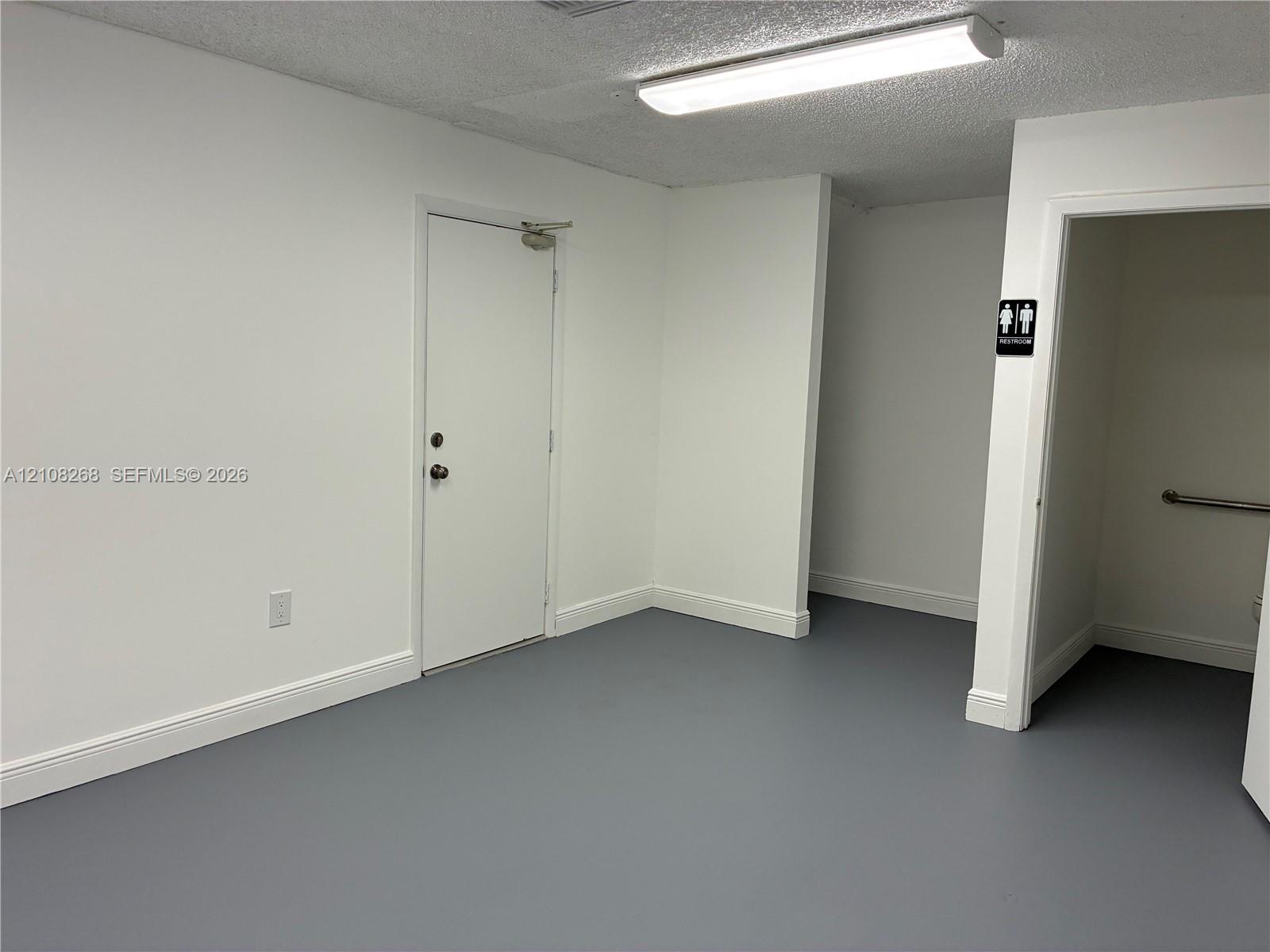 Interior space featuring gray seamless flooring, crisp white walls, a flush-mount rectangular light fixture, and an entry door with commercial-style hardware