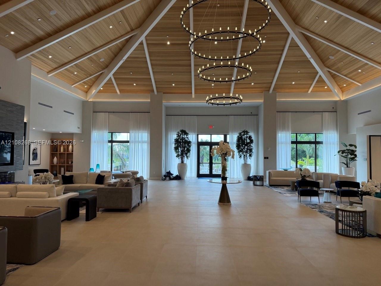 Vaulted wood-plank ceiling featuring exposed beams, stacked three-tier ring chandeliers, large-format tile flooring, and floor-to-ceiling windows