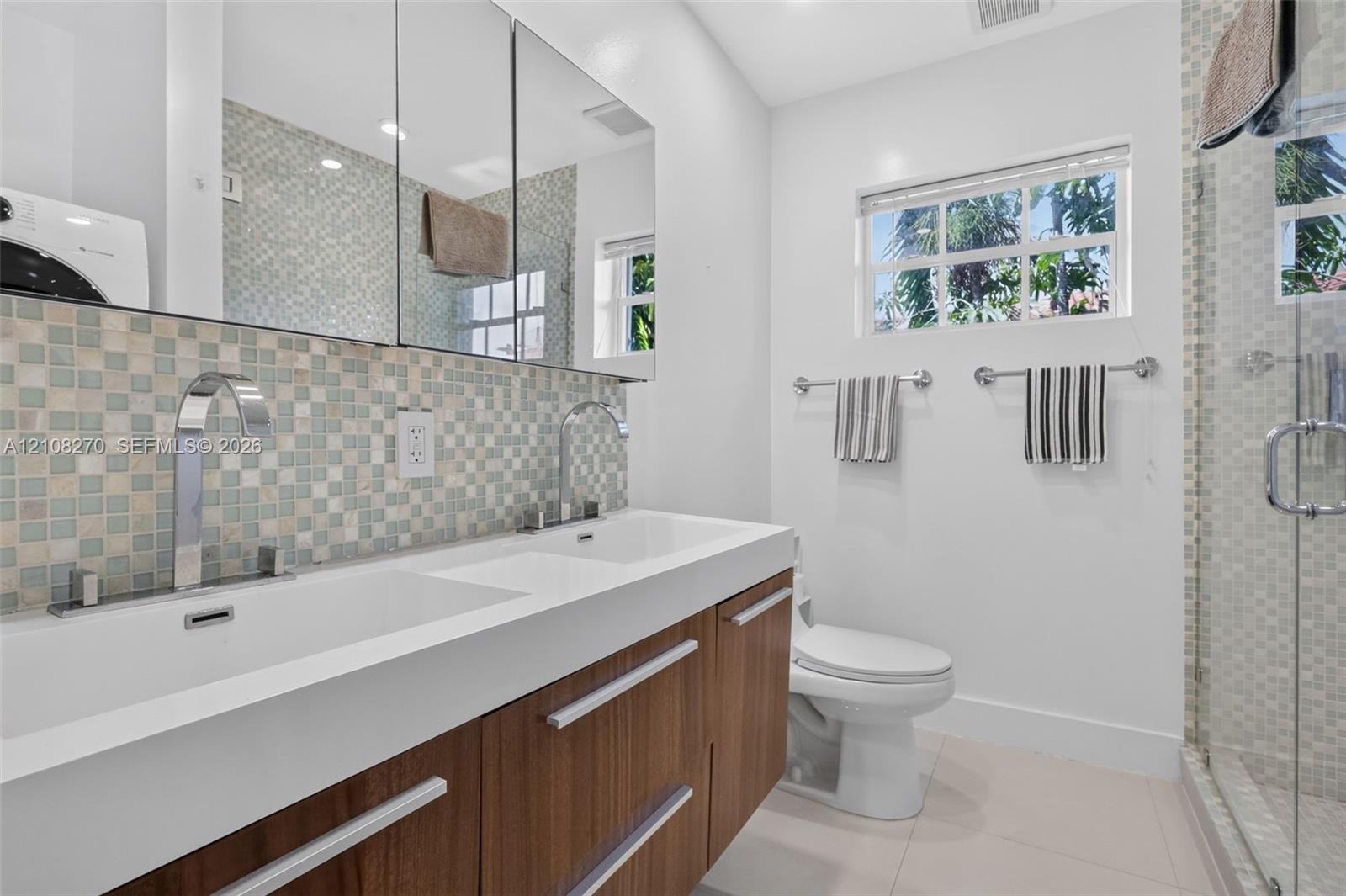 Dual-sink floating vanity with wood-finish cabinetry, mosaic tile backsplash, mirrored medicine cabinets, and glass-enclosed shower