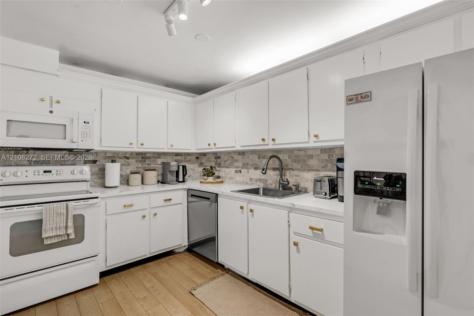 Kitchen featuring white cabinetry with gold-tone hardware, stone-style tile backsplash, white countertops, stainless steel undermount sink with a gooseneck faucet, and wood-finish flooring
