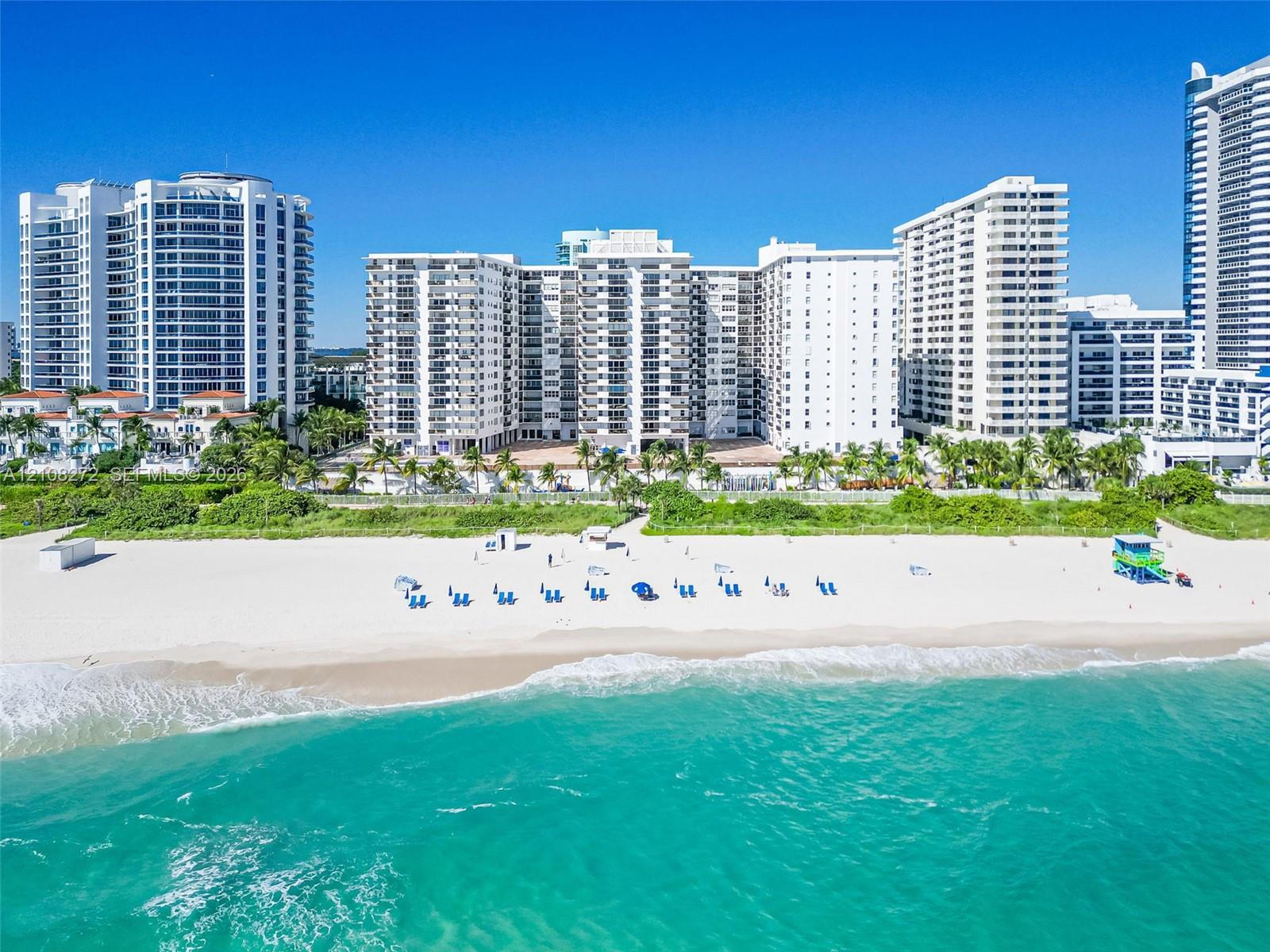 High-rise residential building featuring expansive floor-to-ceiling glass balconies, concrete facade, manicured palm landscaping, and direct access to a sandy beach