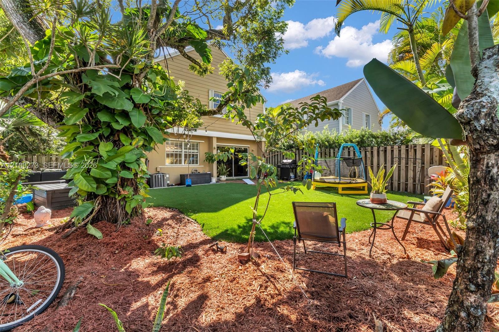 Rear exterior featuring a covered patio, synthetic turf lawn, wood privacy fencing, and mature trees