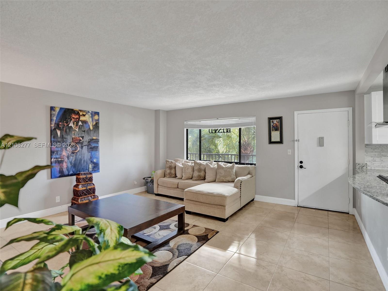 Open-concept interior featuring neutral wall tones, large-format tile flooring, a wide window with blinds, and a stone-surface kitchen counter