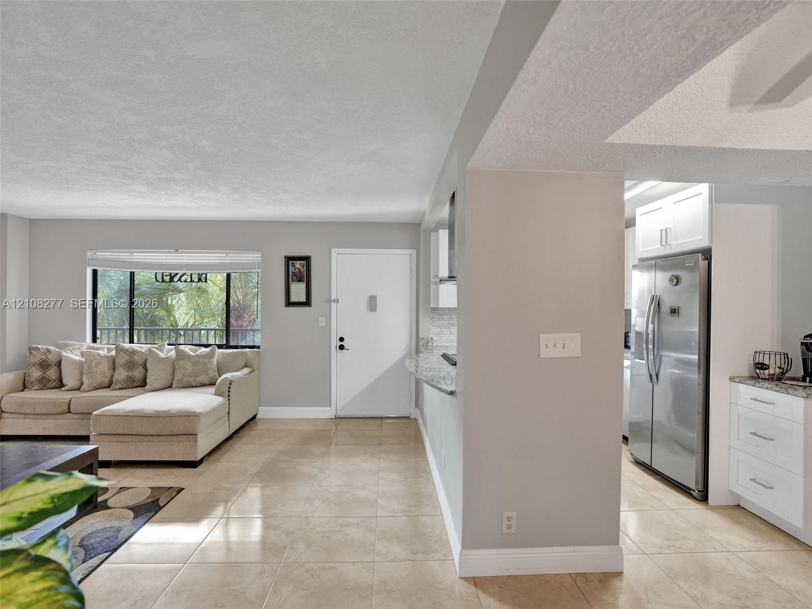 Open-concept interior featuring large-format tile flooring, recessed kitchen cabinetry, stainless steel refrigerator, and oversized window with exterior greenery