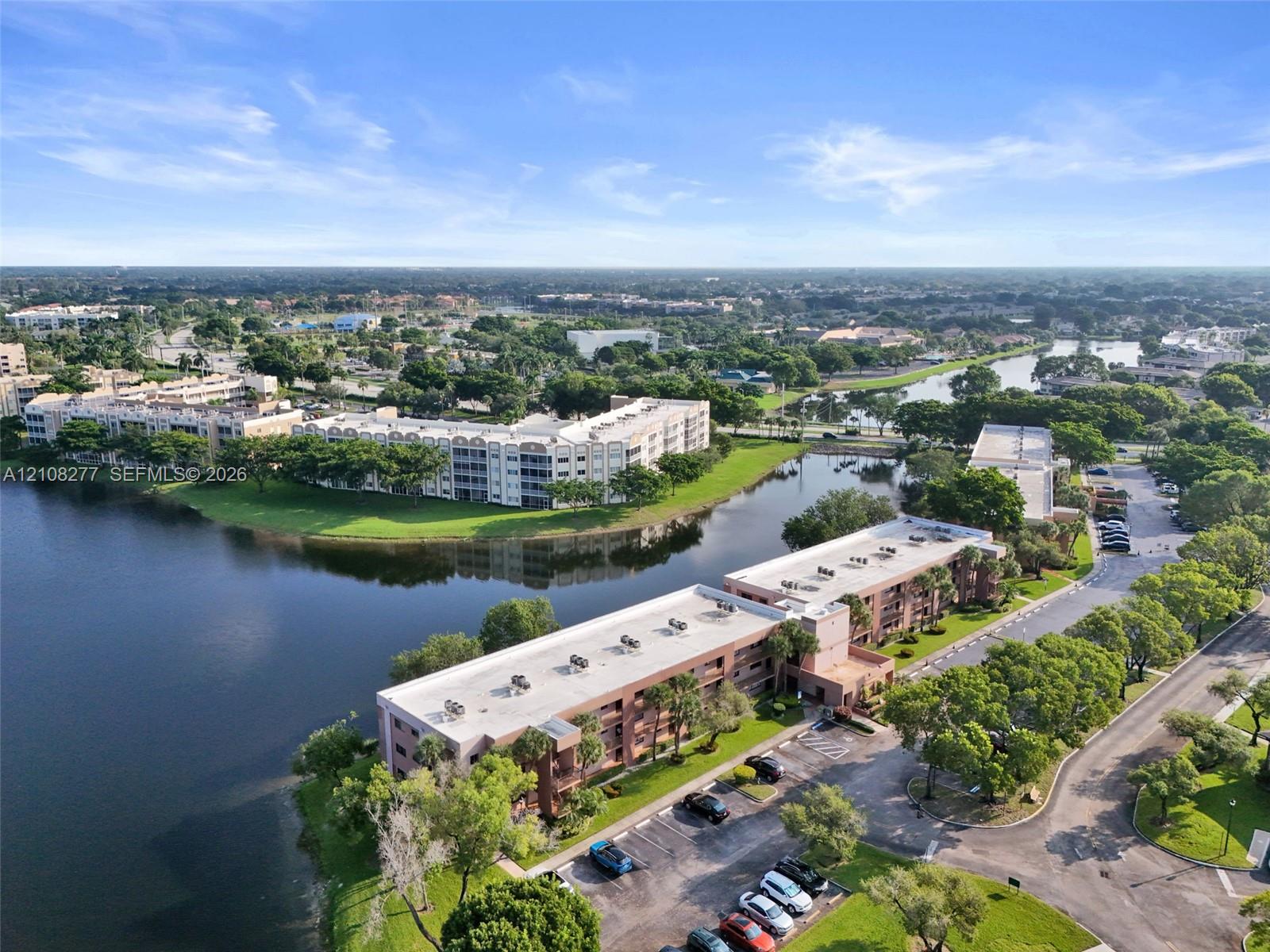 Multi-story residential complex featuring waterfront positioning, expansive exterior parking, manicured landscaping, and mature trees