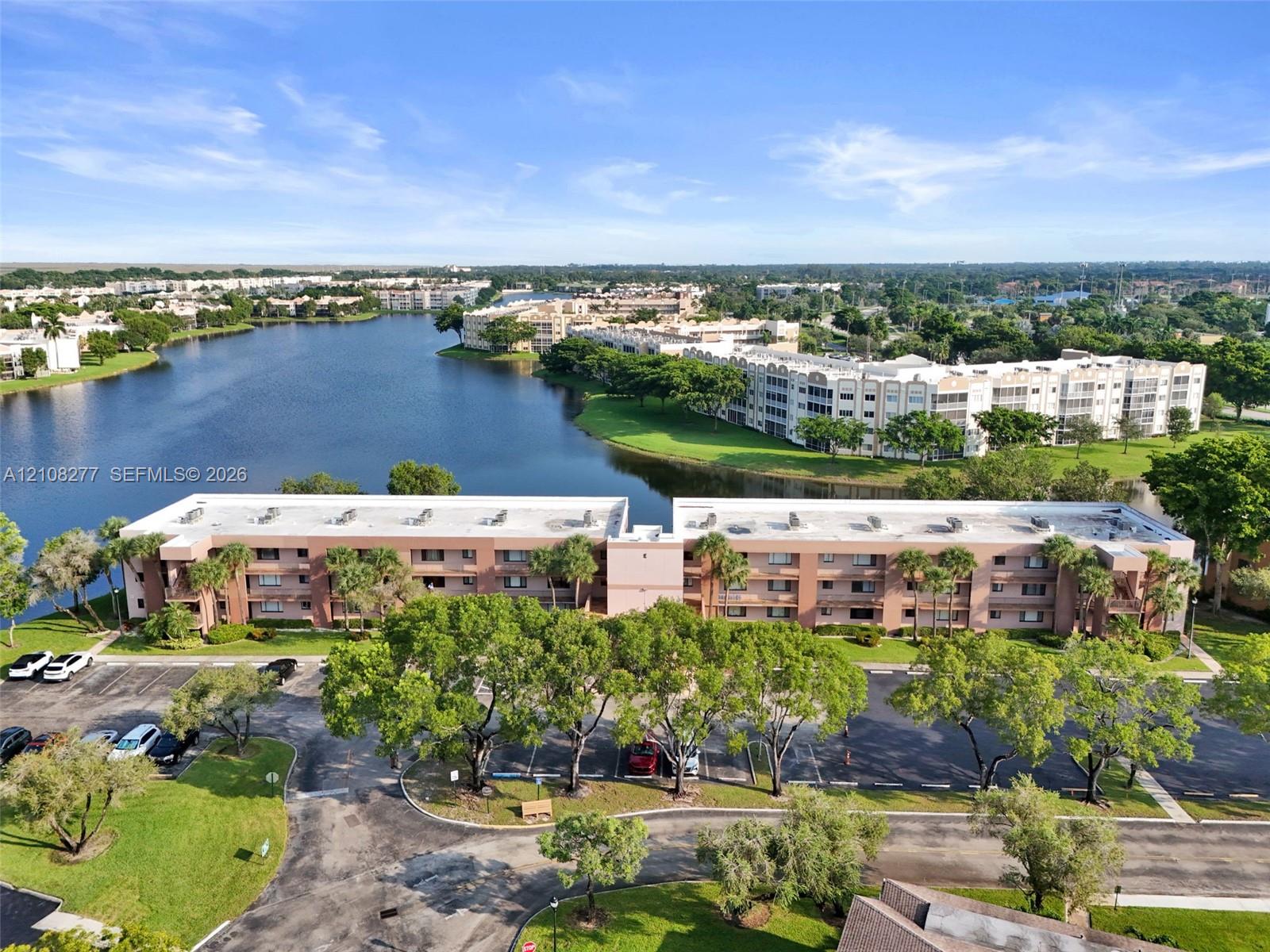 Multi-story residential building featuring stucco exterior, expansive lake frontage, mature trees, and dedicated surface parking