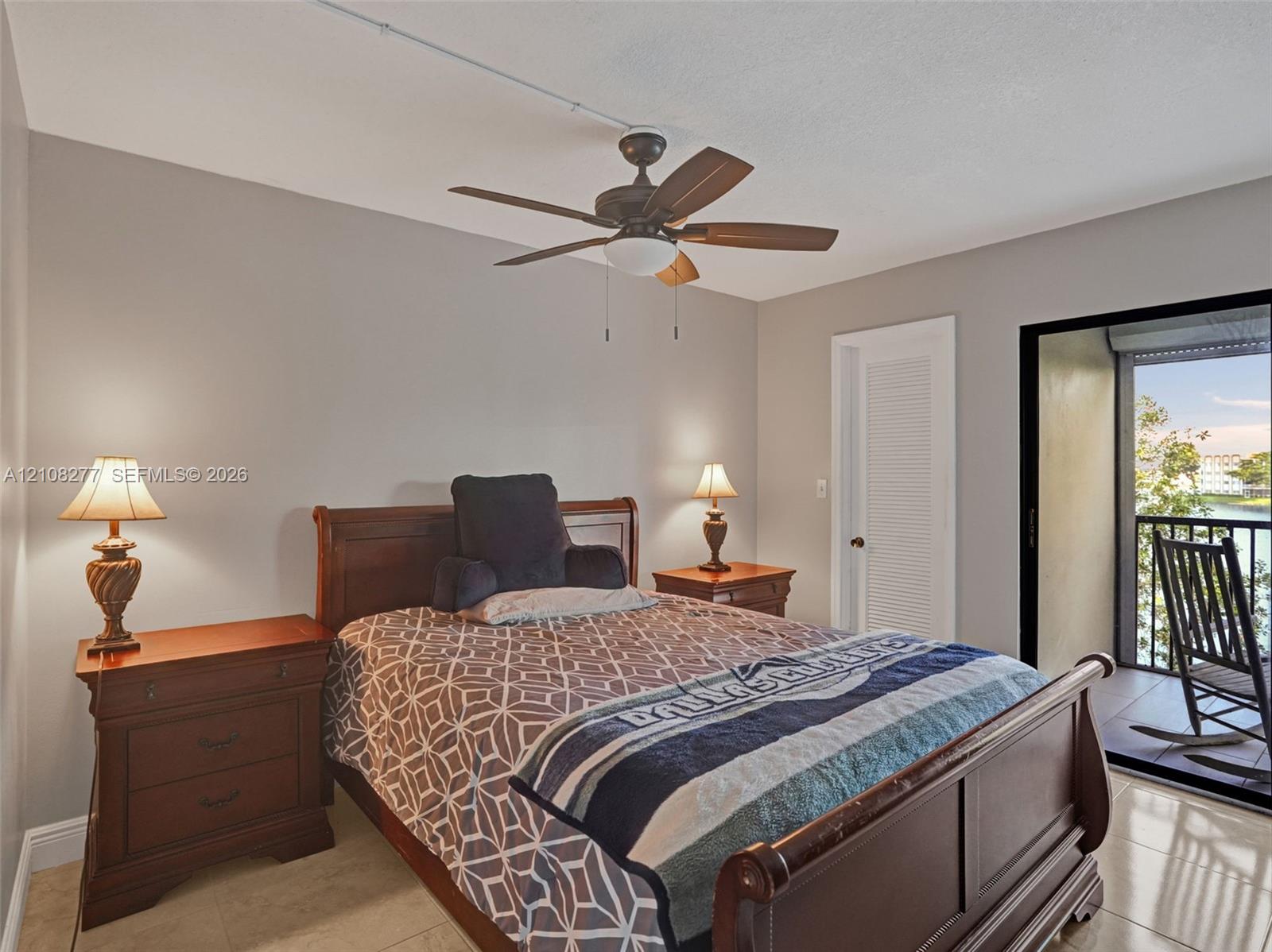 Neutral-toned interior space featuring light-colored tile flooring, a multi-blade ceiling fan, and a louvered closet door with a sliding glass door providing access to a covered balcony