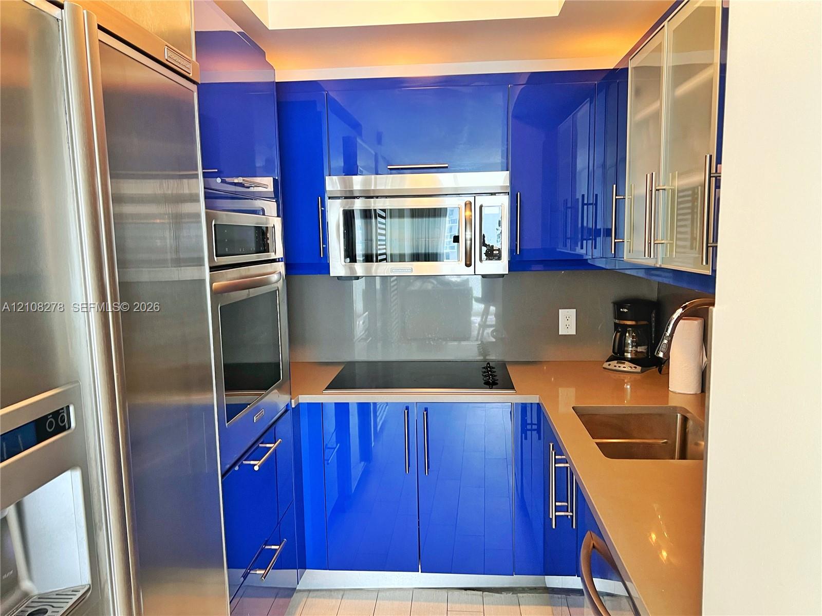 High-gloss blue cabinetry with integrated hardware, neutral solid-surface countertops, stainless steel wall-mounted oven and microwave, professional-grade refrigerator, and a stainless steel undermount sink
