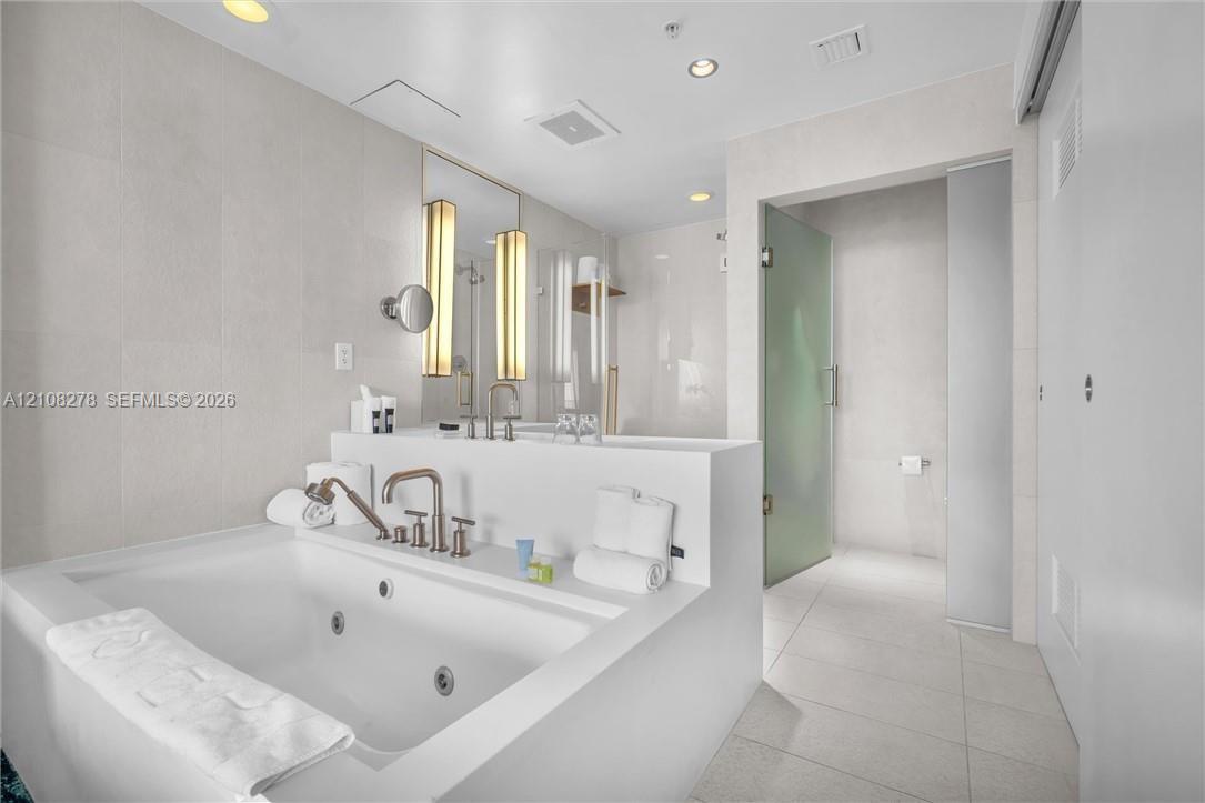 Jetted soaking tub with deck-mounted metallic fixtures, large-format tile wall surround, dual integrated vanity sinks with sconce lighting, and a walk-in glass-enclosed shower