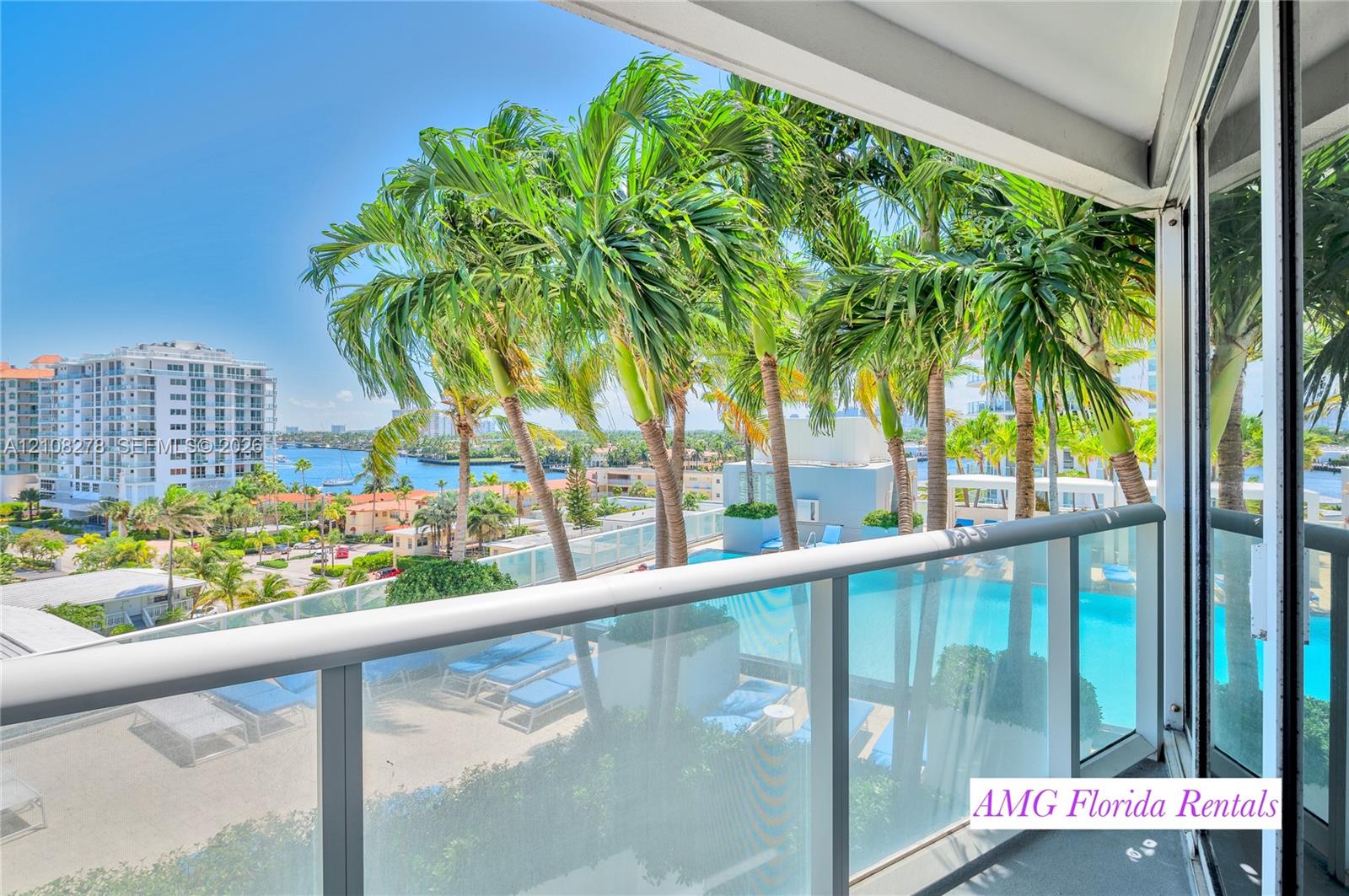 Private balcony with glass panel railing overlooking a swimming pool, mature palm trees, and coastal waterway