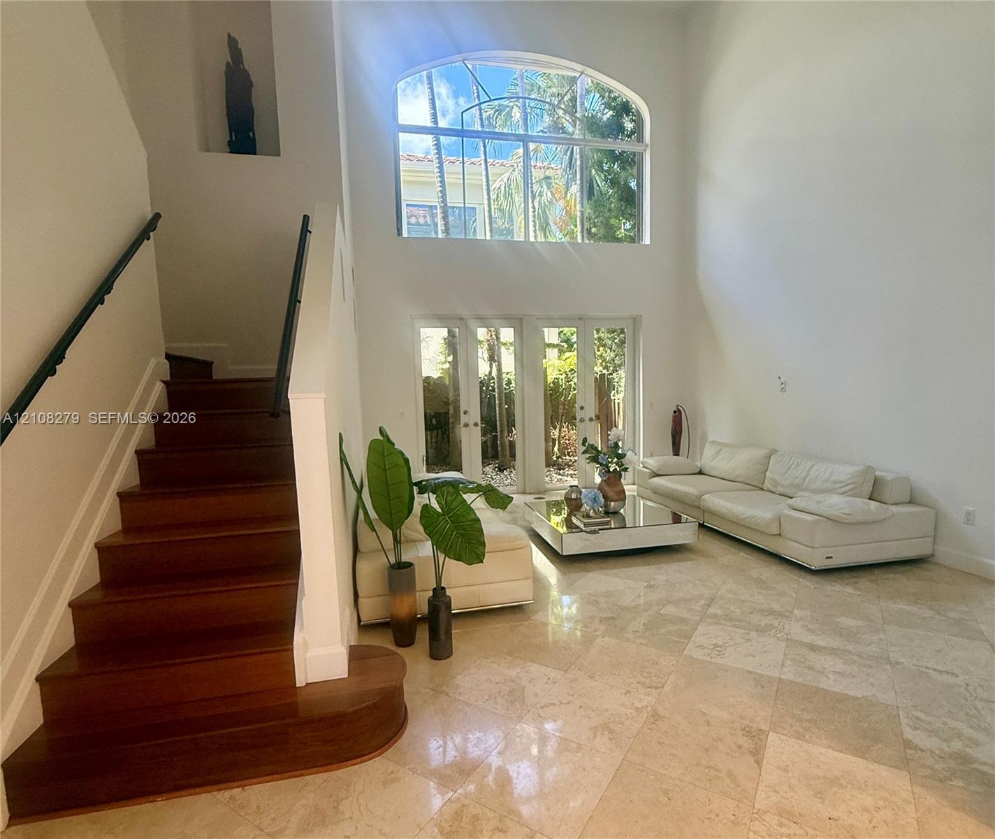 High-ceiling interior featuring stone tile flooring, a wood-finish staircase with a wrought-iron handrail, a large arched window, and full-height glass-paned doors
