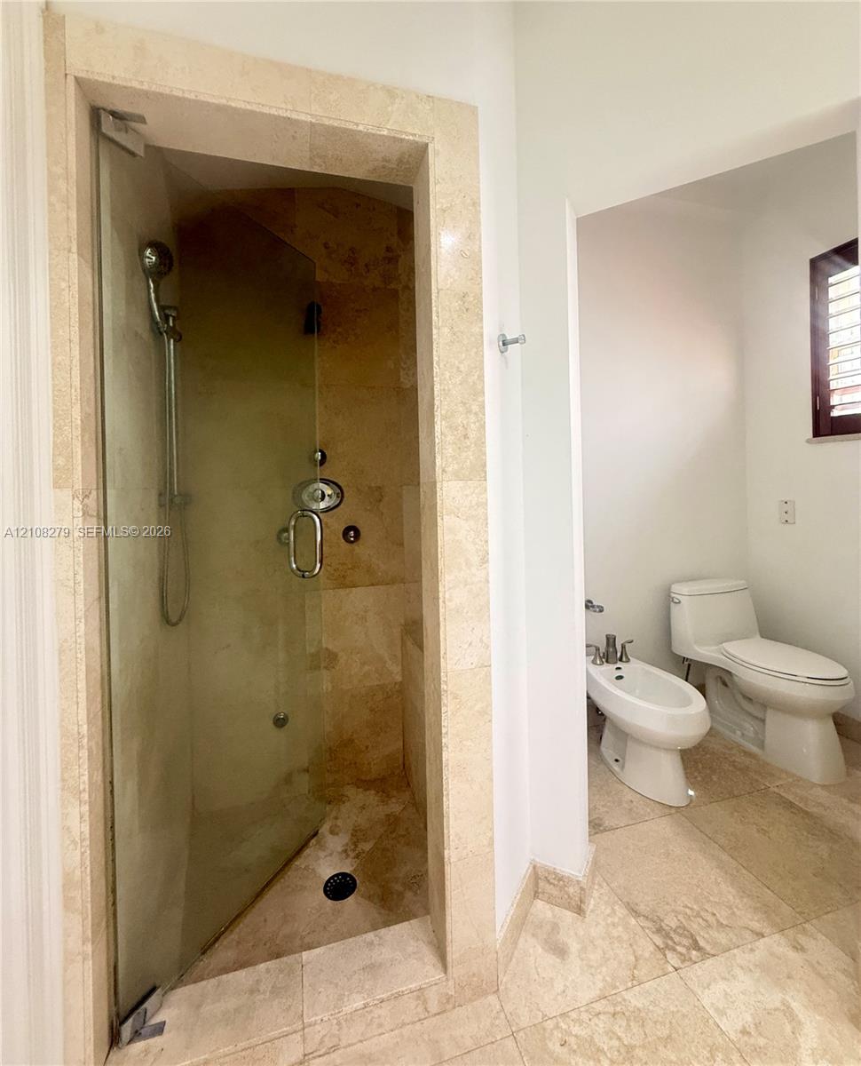 Walk-in shower featuring a frameless glass door, stone-tile surround, and handheld showerhead, accompanied by a bidet and toilet with stone-tile flooring