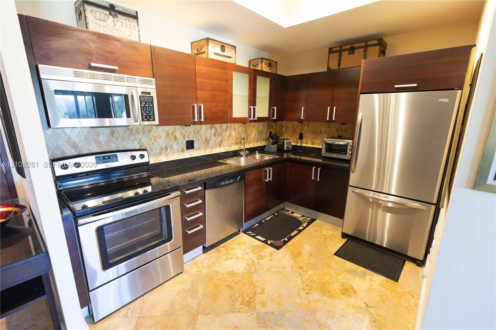 Kitchen featuring dark wood-finish cabinetry, stainless steel appliances, stone-finish countertops, geometric-pattern backsplash tile, and stone-finish flooring