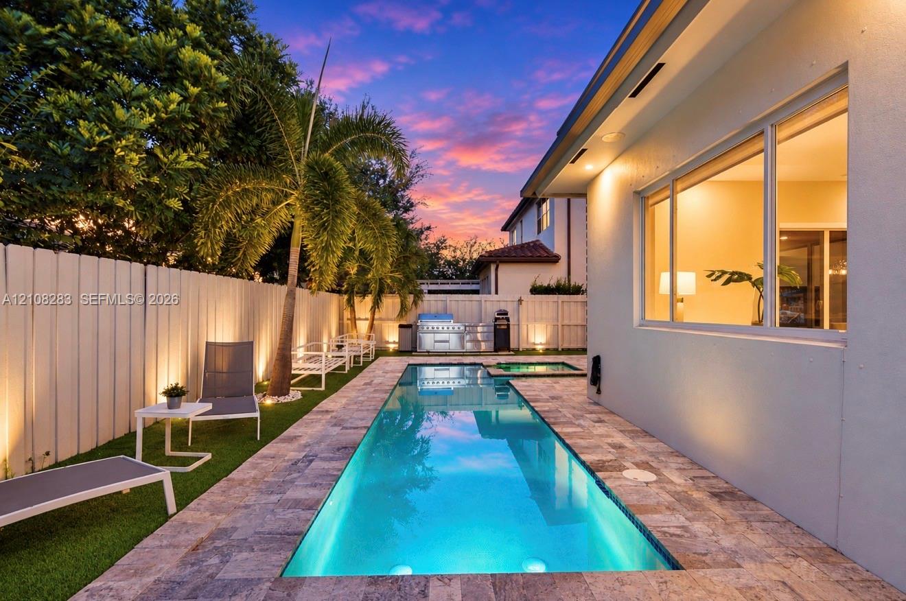 Rectangular swimming pool featuring a stone-paver surround, integrated spa, built-in outdoor grill station, and artificial turf lawn, enclosed by privacy fencing