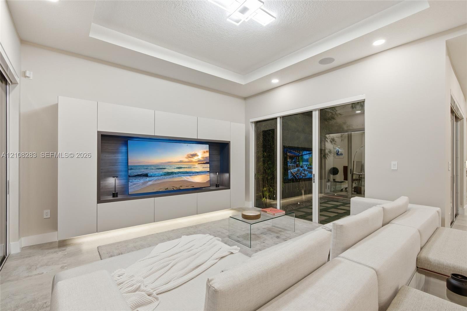 Living area featuring a tray ceiling with recessed lighting, custom floor-to-ceiling built-in media cabinetry with integrated lower illumination, and glass sliding doors