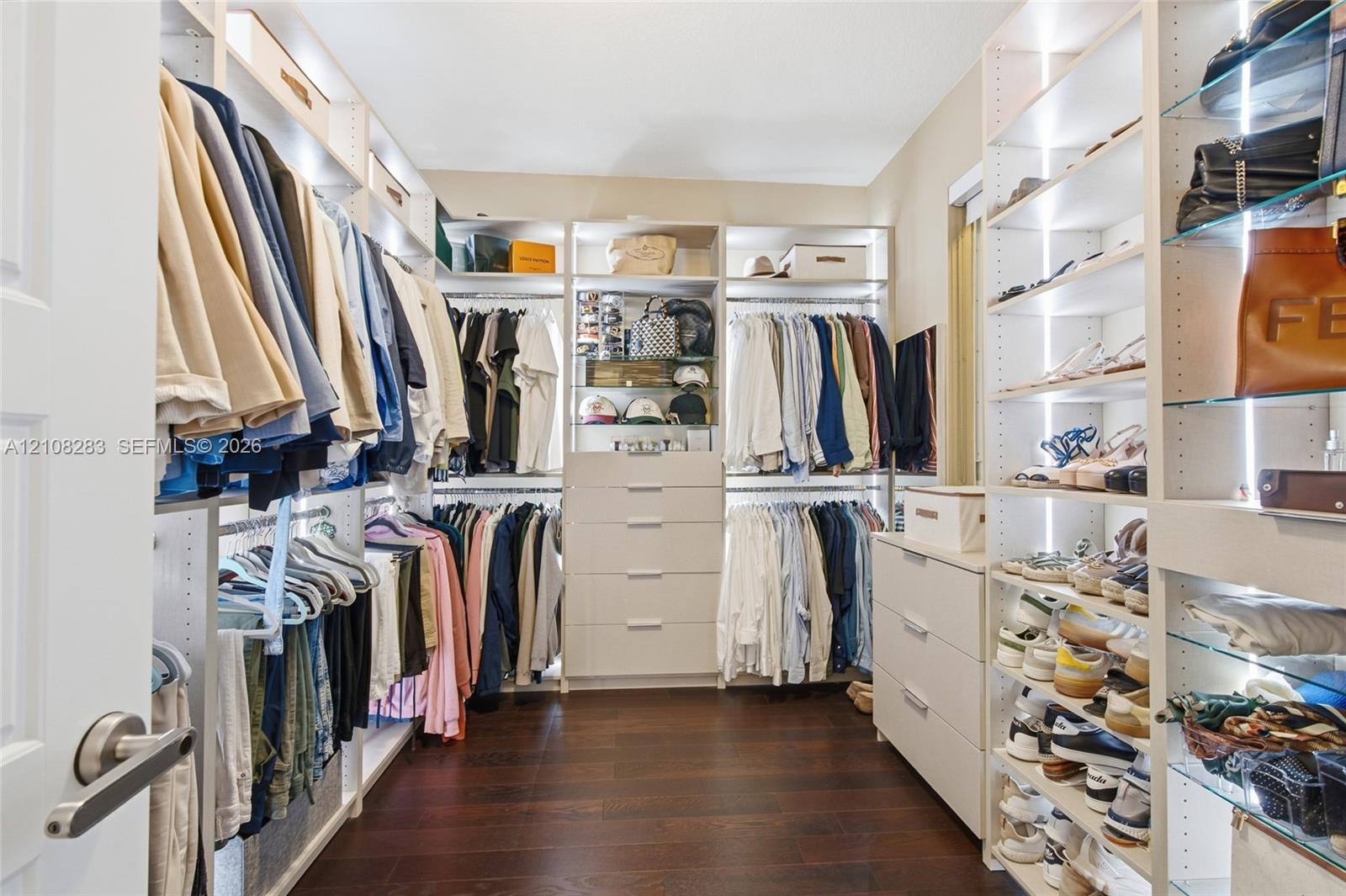 Walk-in closet featuring floor-to-ceiling custom built-in shelving, glass display shelving with integrated lighting, multi-tiered hanging rods, and integrated drawer units, all set upon dark wood-finish flooring
