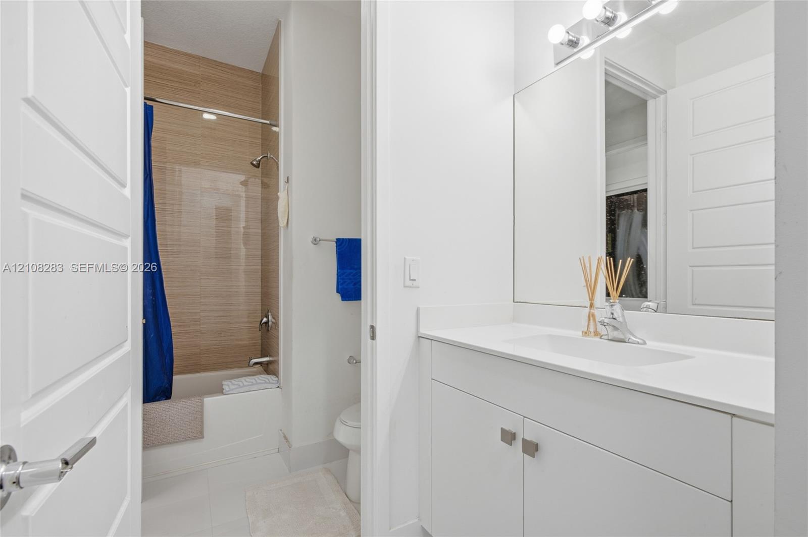 Bathroom featuring a white vanity with a composite countertop, an integrated rectangular sink, a floor-to-ceiling tiled shower surround with a light wood-finish pattern, and a wall-mounted light fixture above a large mirror