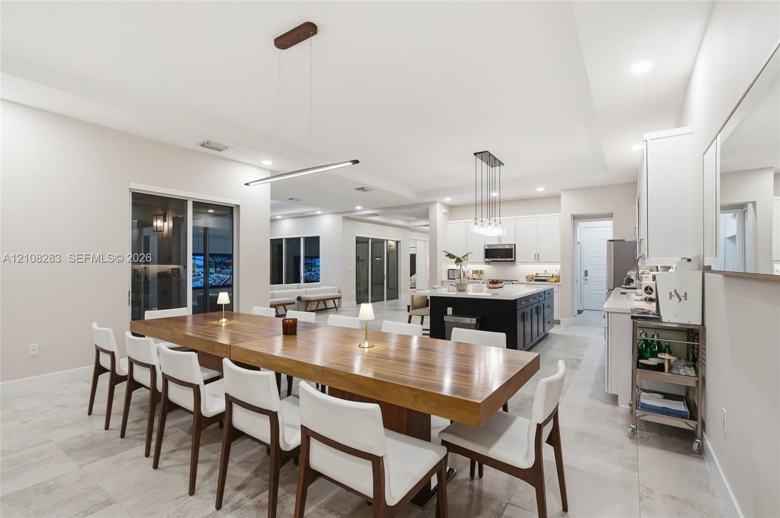 Open concept kitchen and dining area featuring large format tile flooring, a central kitchen island with contrasting cabinetry, white shaker-style wall cabinets, and recessed lighting