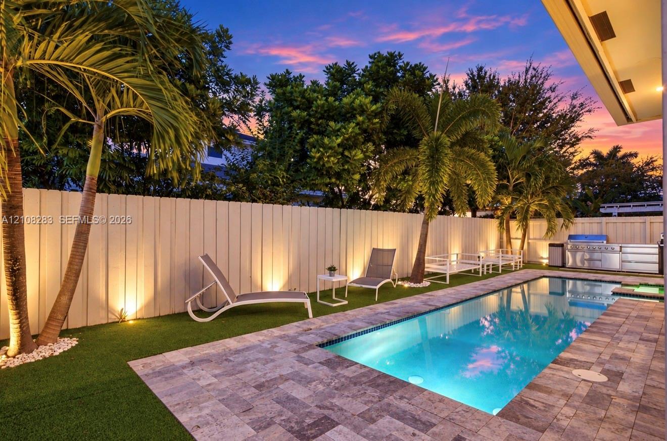 Backyard featuring a rectangular swimming pool, stone-paver decking, integrated outdoor kitchen, artificial turf, and perimeter wall lighting