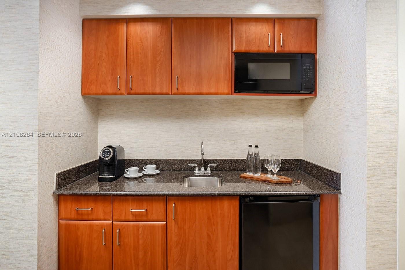 Wet bar featuring wood-finish cabinetry, stone countertop, stainless steel drop-in sink, and built-in microwave